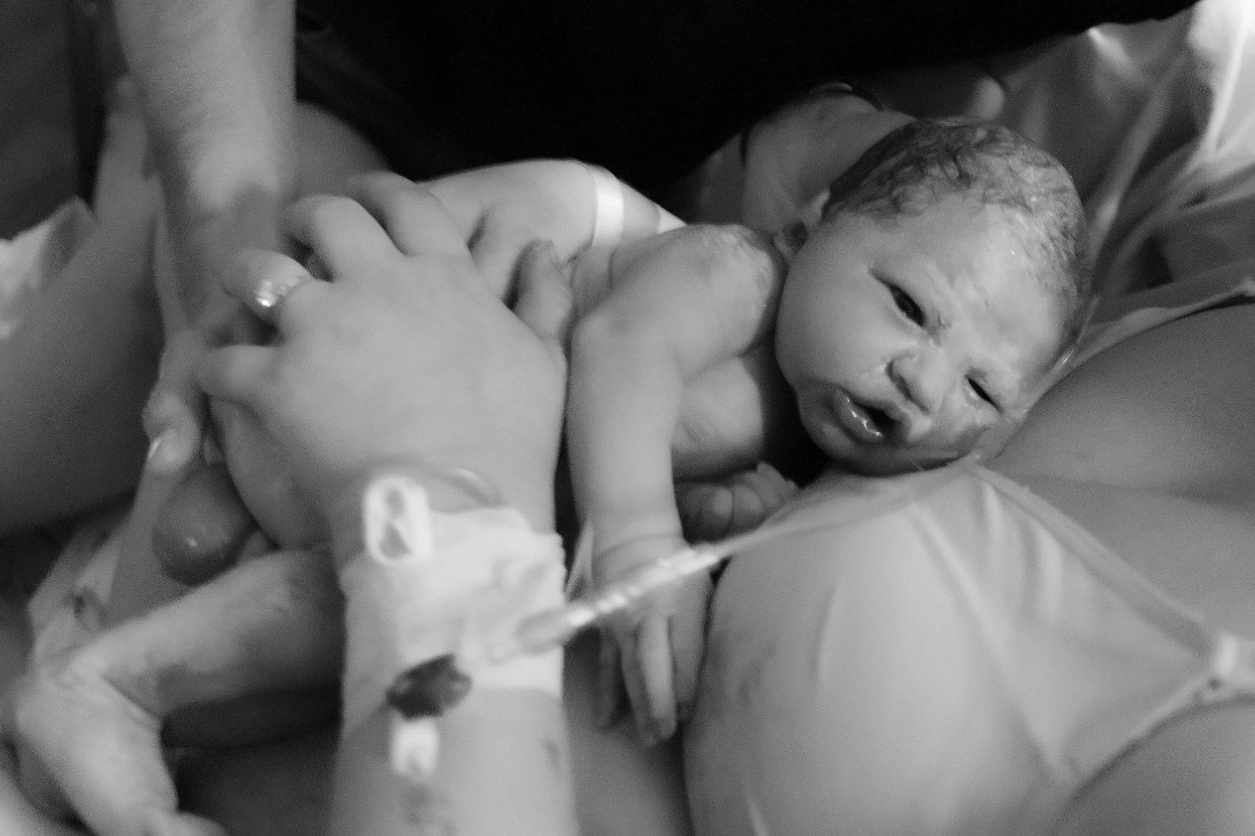 Newborn baby being held by medical staff, lying on an adult's arm, with hospital wristbands visible.