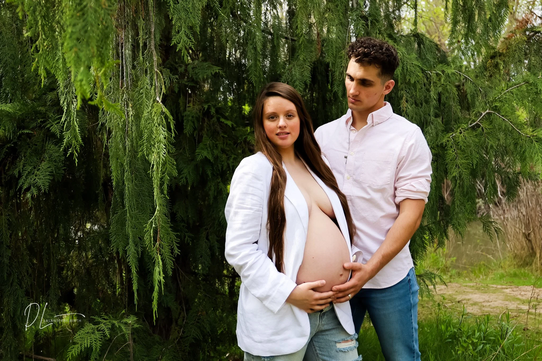 A pregnant woman with long brown hair wearing a white blazer and blue jeans stands outdoors next to a man in a white shirt and blue jeans; they are in front of green foliage. The woman smiles slightly, and the man looks at her with affection, gently