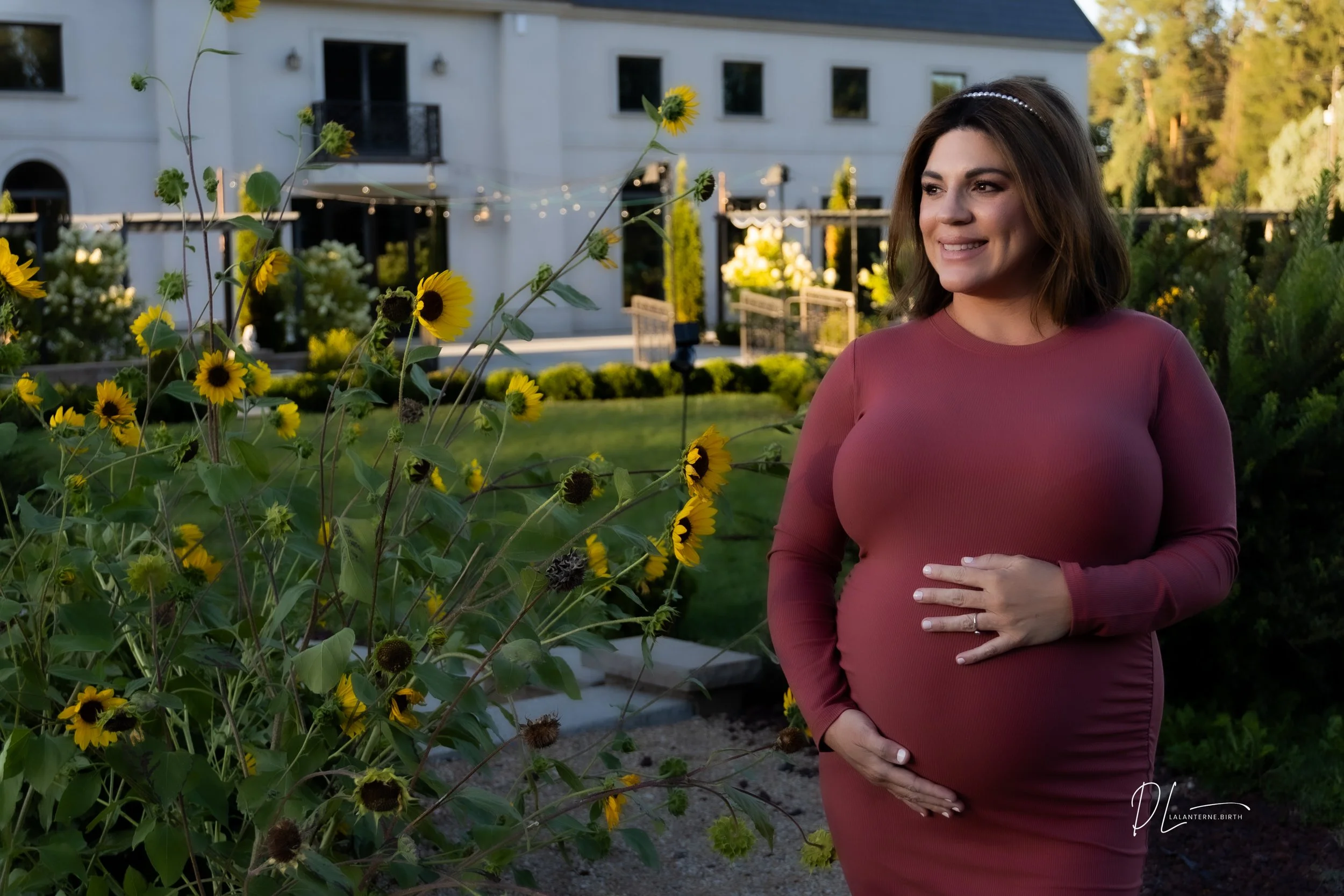 A pregnant woman with shoulder-length brown hair, wearing a long-sleeved mauve dress and a pearl headband, standing outdoors in a garden with yellow sunflowers, smiling and touching her belly with both hands, in the late afternoon or early evening.