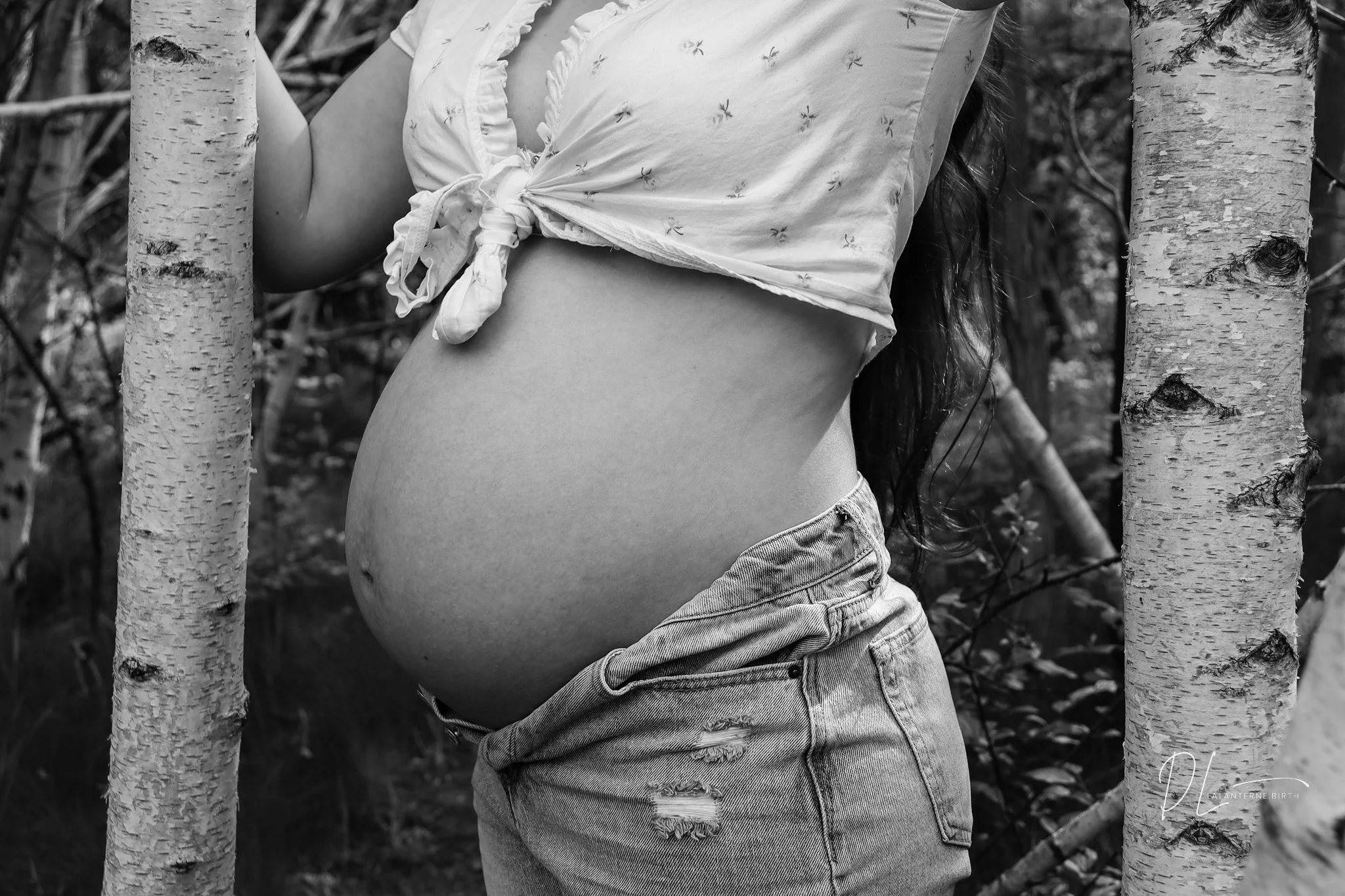A pregnant woman in a knotted white top and ripped jeans stands between two tree trunks in a forest.
