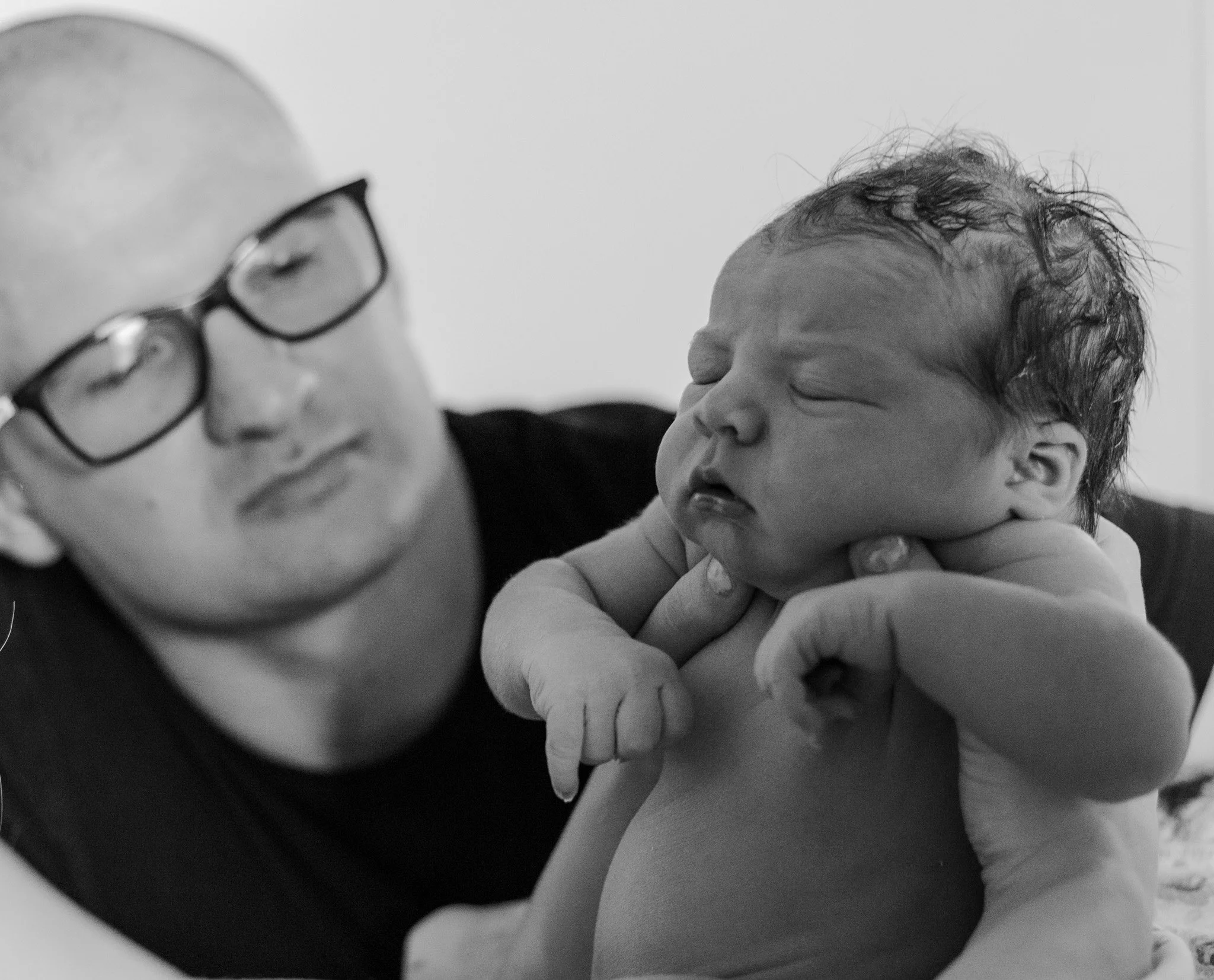 A man wearing glasses holds a sleeping newborn baby up, supporting the baby’s neck and shoulder with his hand in a close-up black and white photograph.