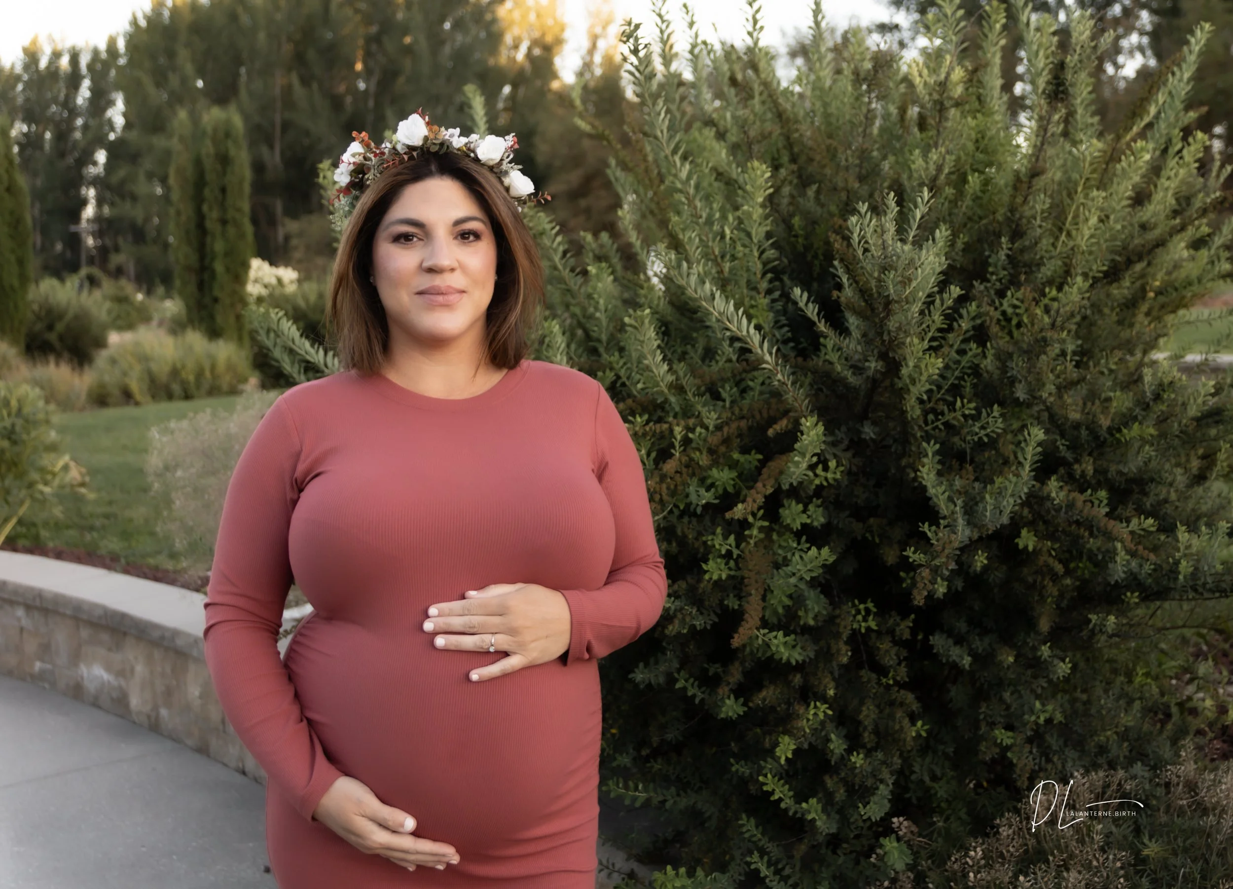 A pregnant woman with shoulder-length brown hair wearing a long-sleeve pink dress and a floral crown standing outdoors near a large bush and trees during sunset, gently holding her baby bump.