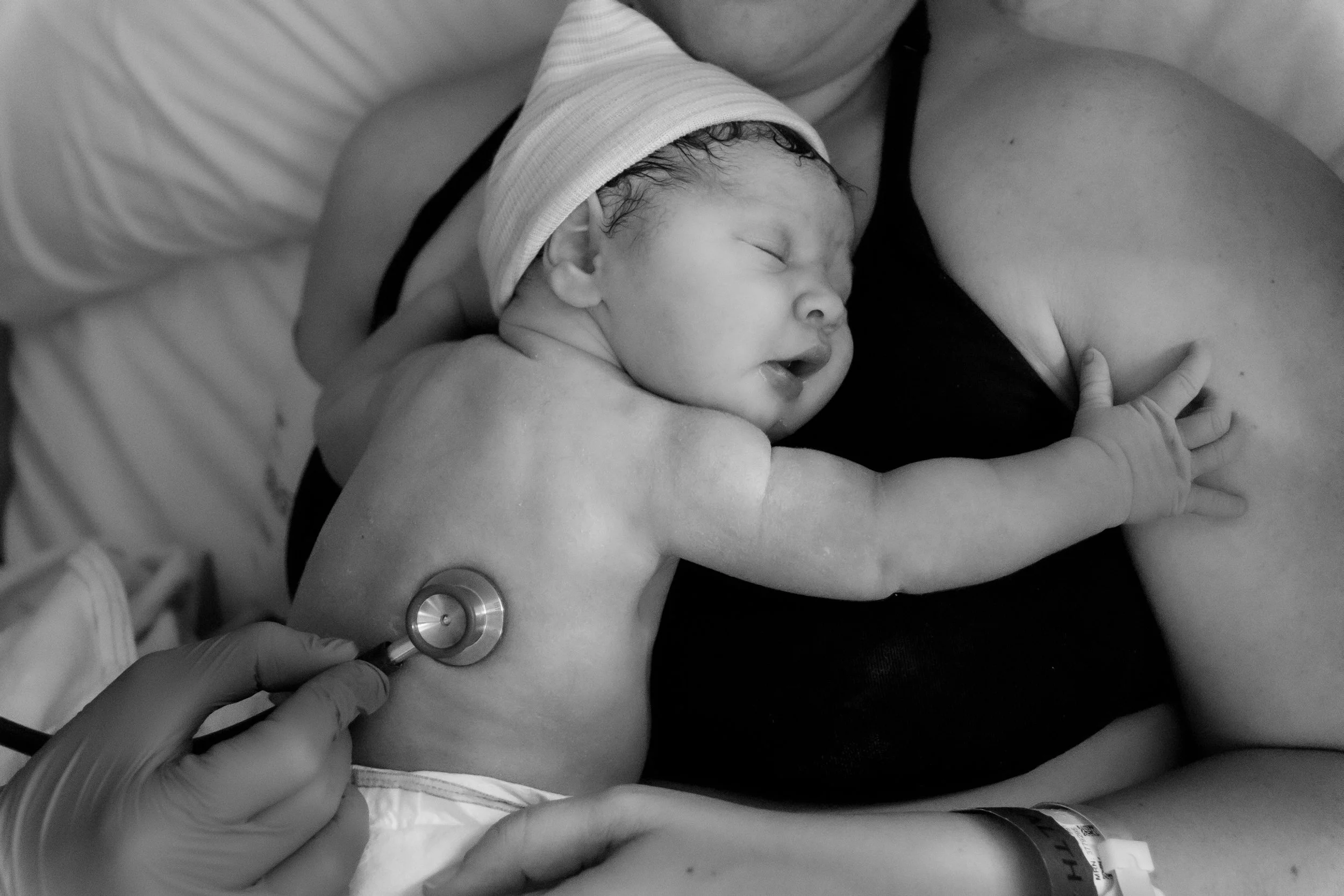 A newborn baby with a cap being examined with a stethoscope on the mother's chest in a hospital room.