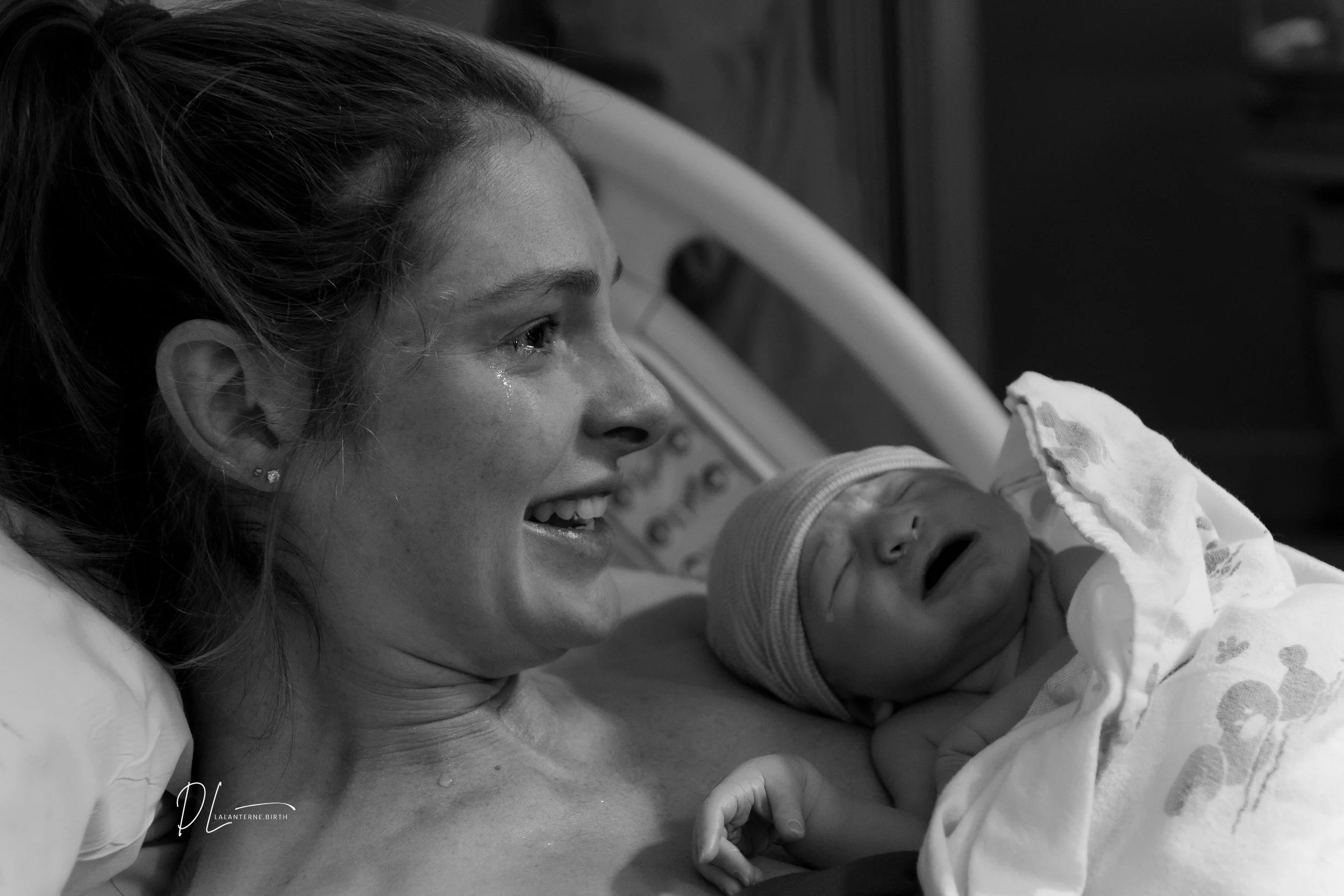 A woman in a hospital bed smiling with tears of joy, holding a newborn baby who is crying, both in a black and white photo.