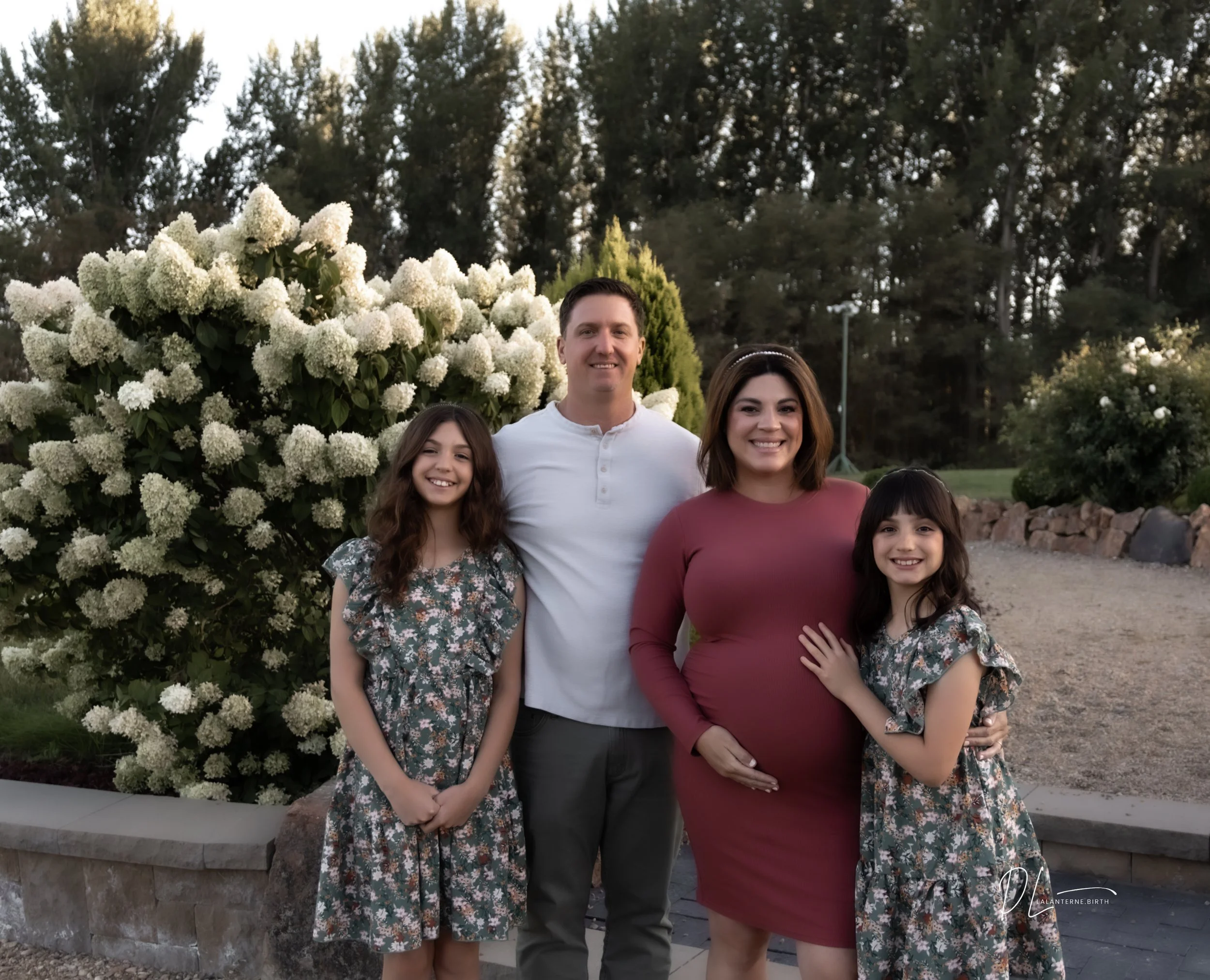 A family of four standing outdoors in front of flowering bushes and trees, smiling at the camera. The woman is pregnant, wearing a maroon dress, and the man is wearing a white shirt. Two young girls in floral dresses are standing on either side.