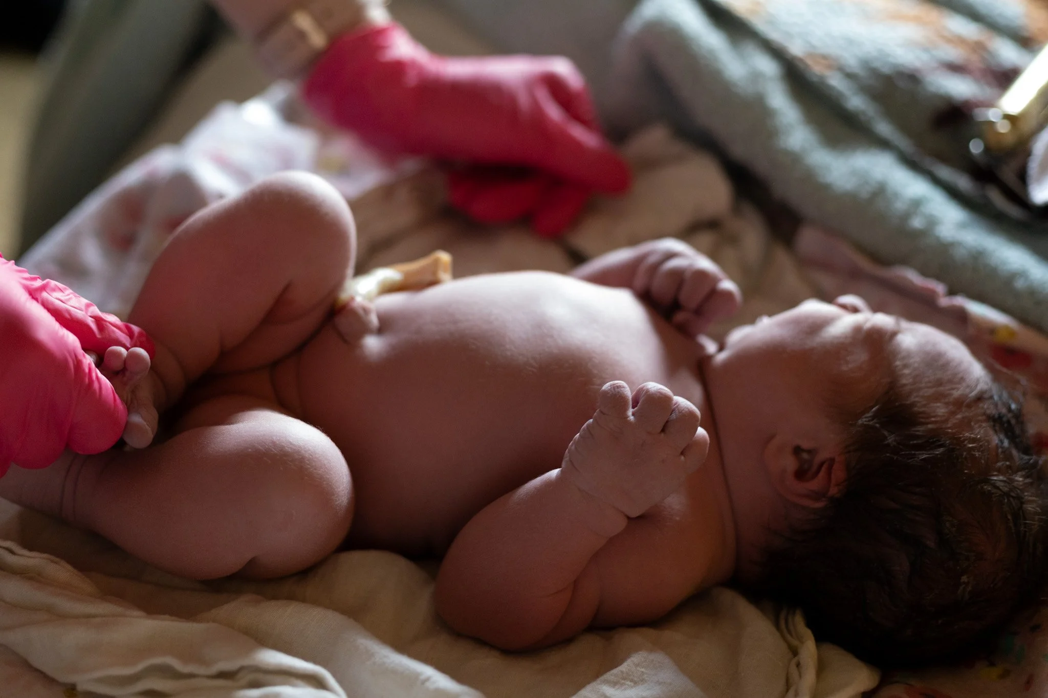 A newborn baby lying on a bed, with a person wearing pink gloves tending to them.