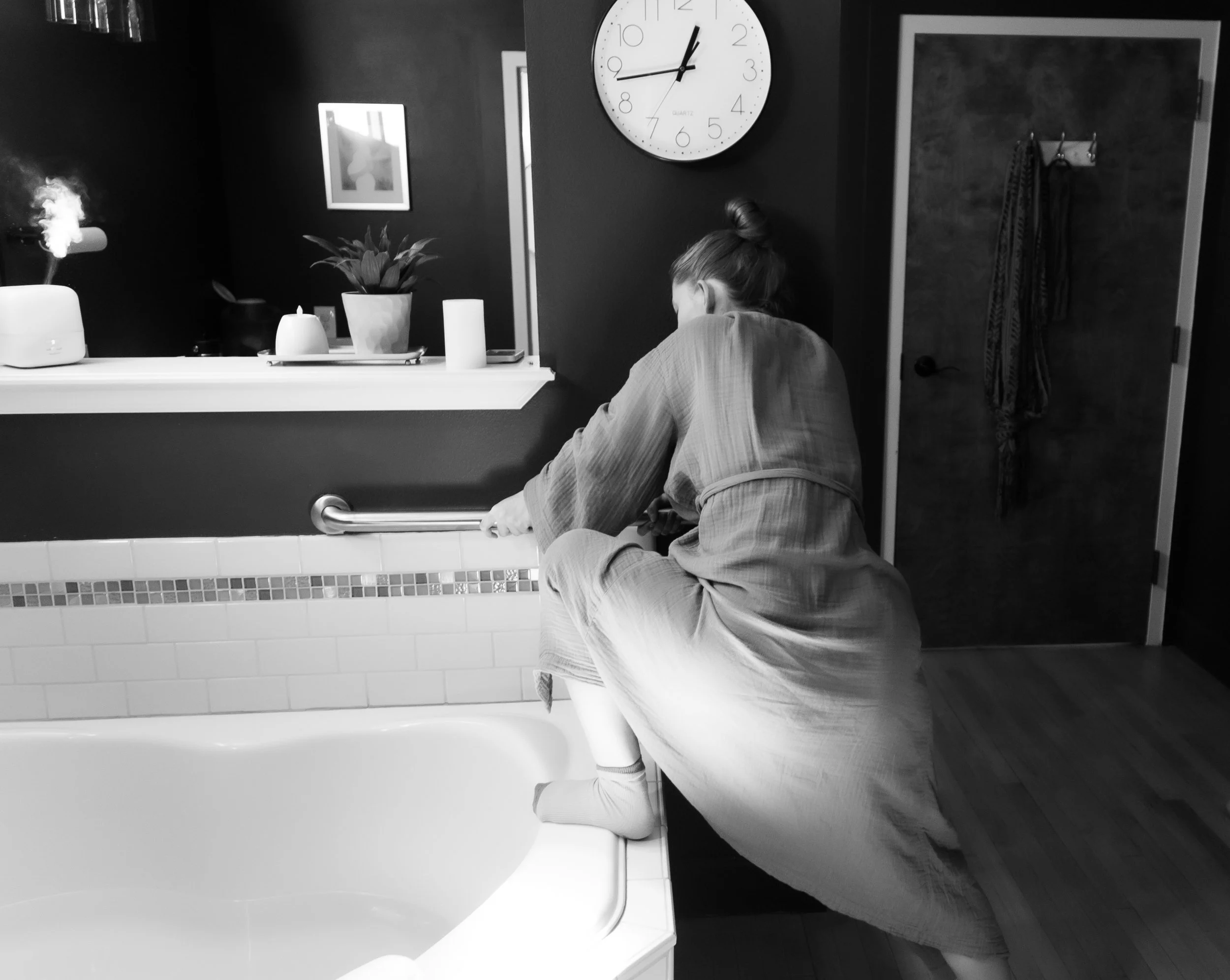 A person dressed in a robe and socks is using a grab bar to steady themselves while stepping into a bathtub in a bathroom.