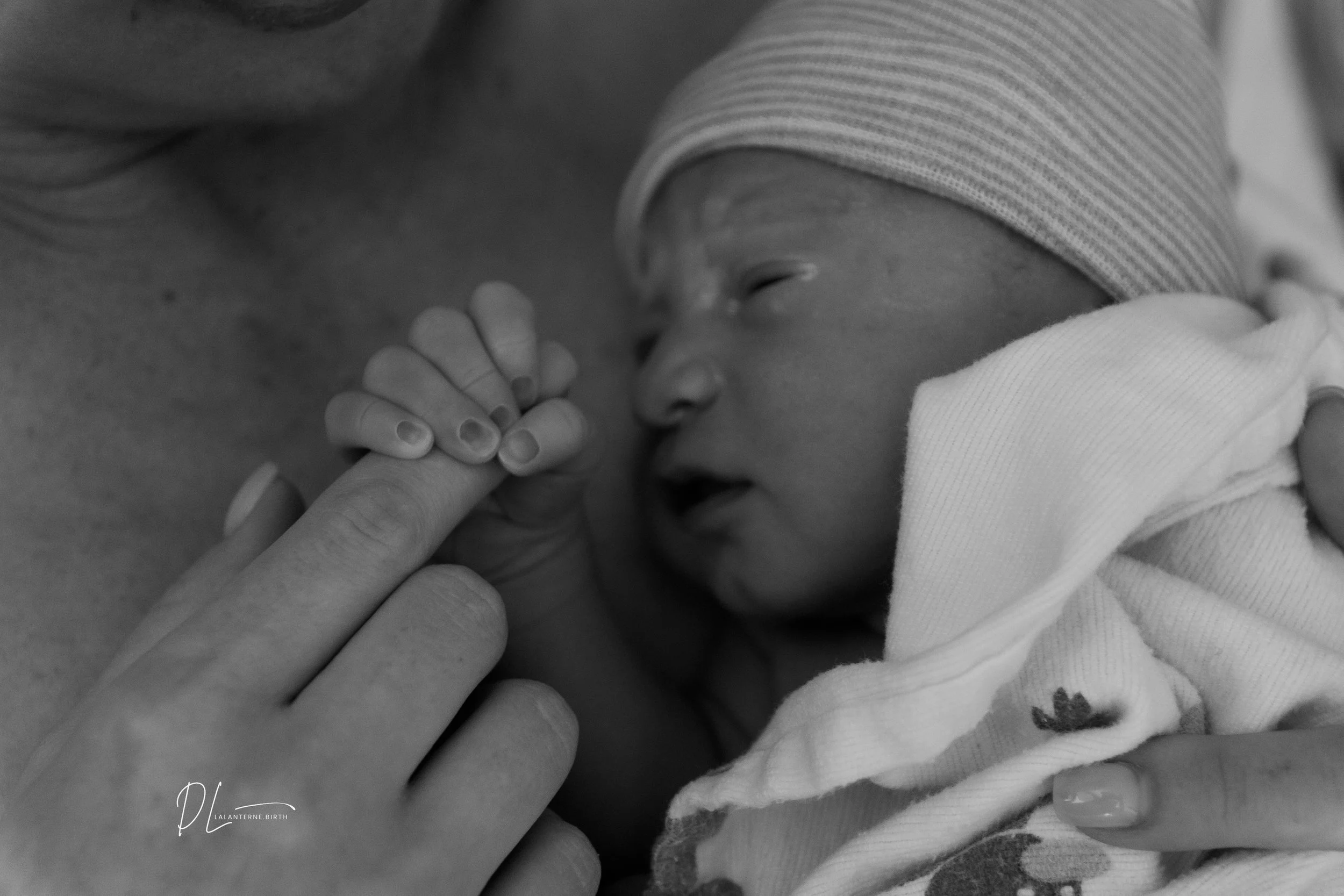 A newborn baby holding an adult's finger, lying on their chest, with eyes closed and wearing a striped cap and a soft blanket.