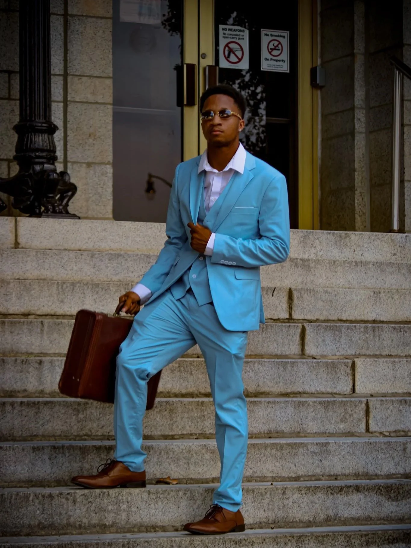 A young man in a light blue suit, white shirt, and brown dress shoes standing on stone stairs in front of a building with glass doors. He is holding a brown briefcase and wearing sunglasses. Fayetteville, NC