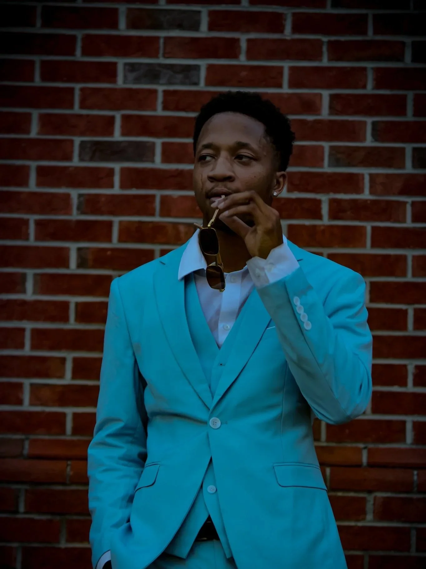 Young man in a light blue suit leaning against a brick wall, holding sunglasses and looking thoughtfully to the side. Fayetteville, NC