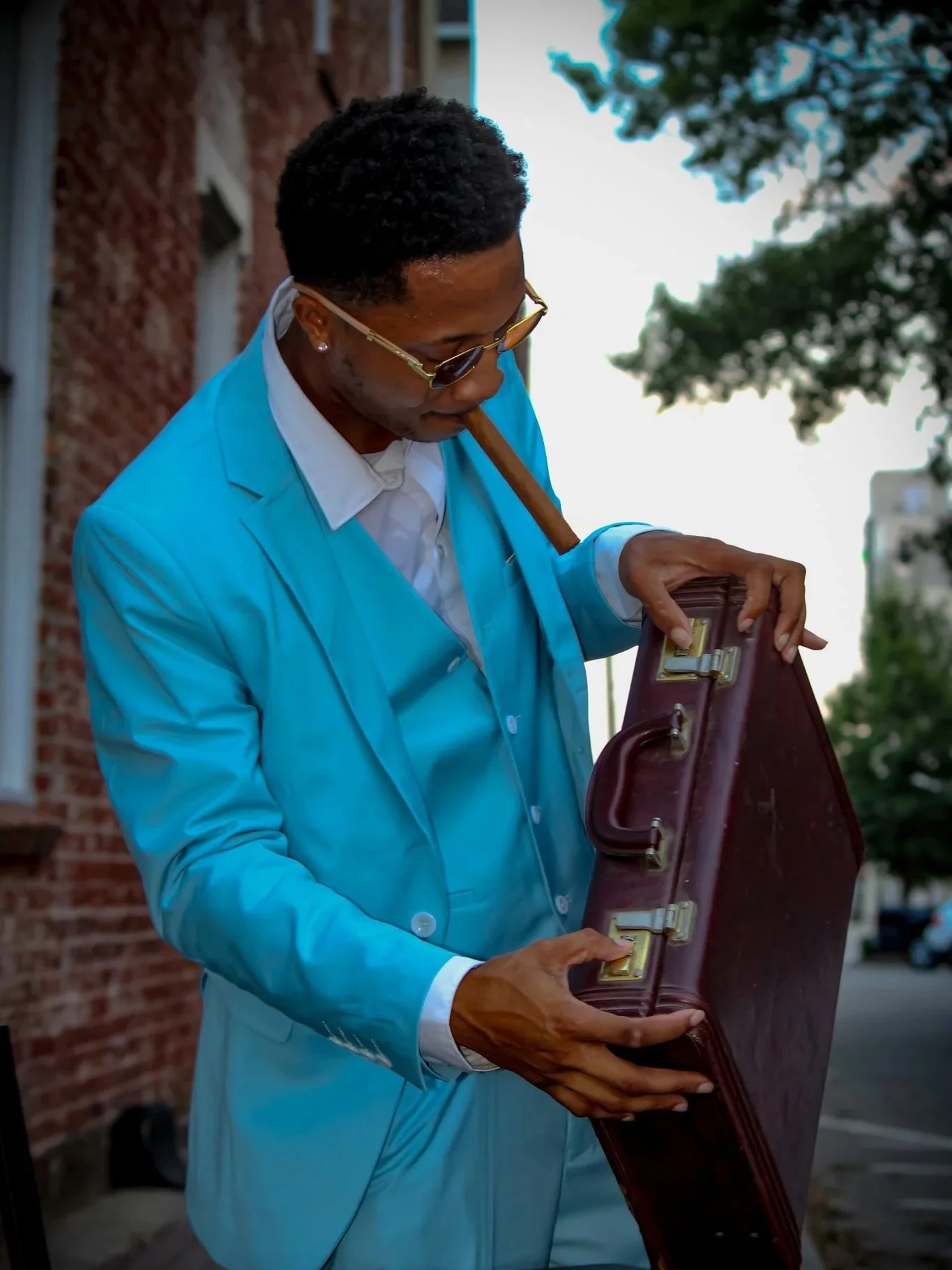 A man in a bright blue suit with sunglasses holding a briefcase outdoors in front of a brick building with trees in the background. 
Fayetteville, NC