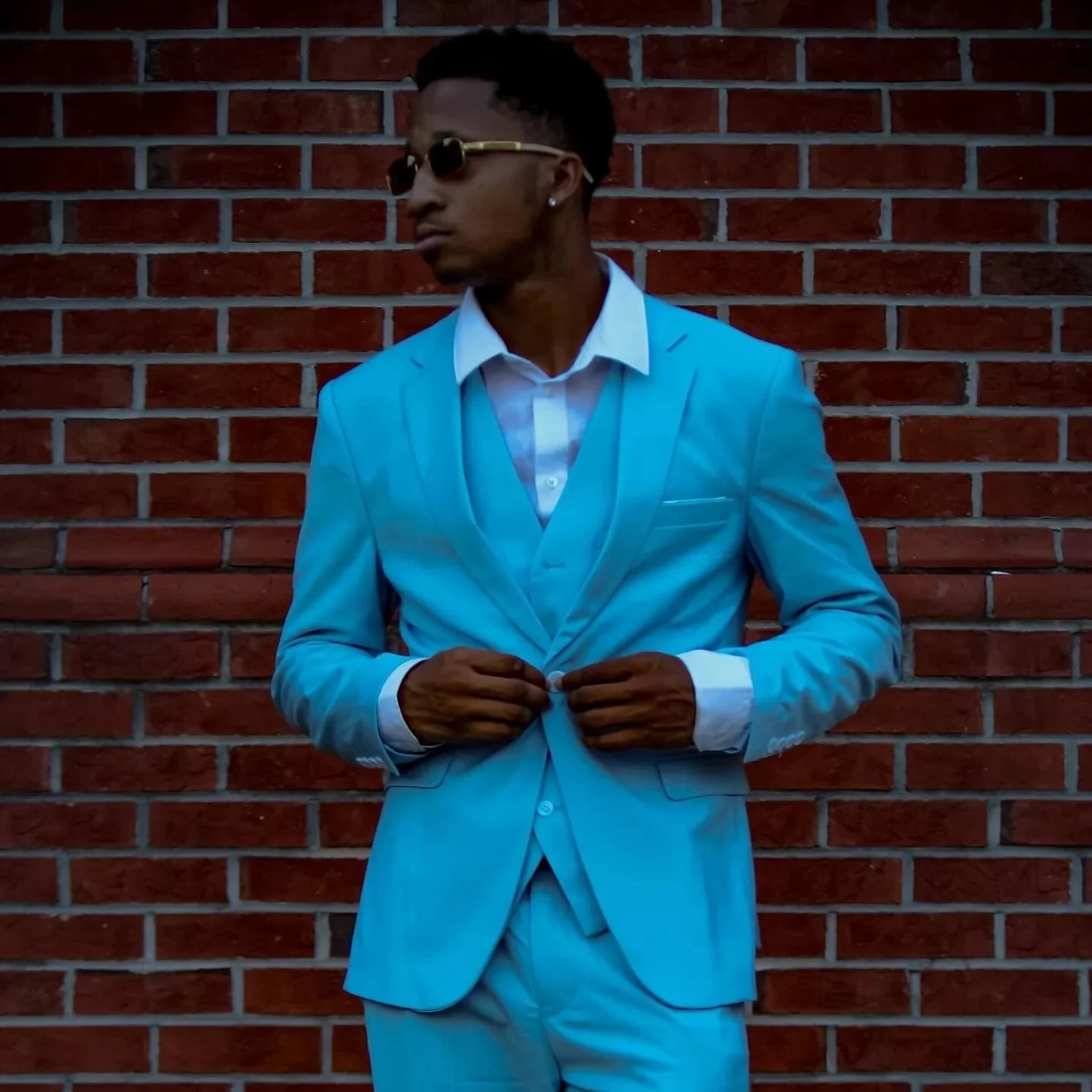 A man wearing a light blue suit, white shirt, and sunglasses standing against a red brick wall. Fayetteville, NC
