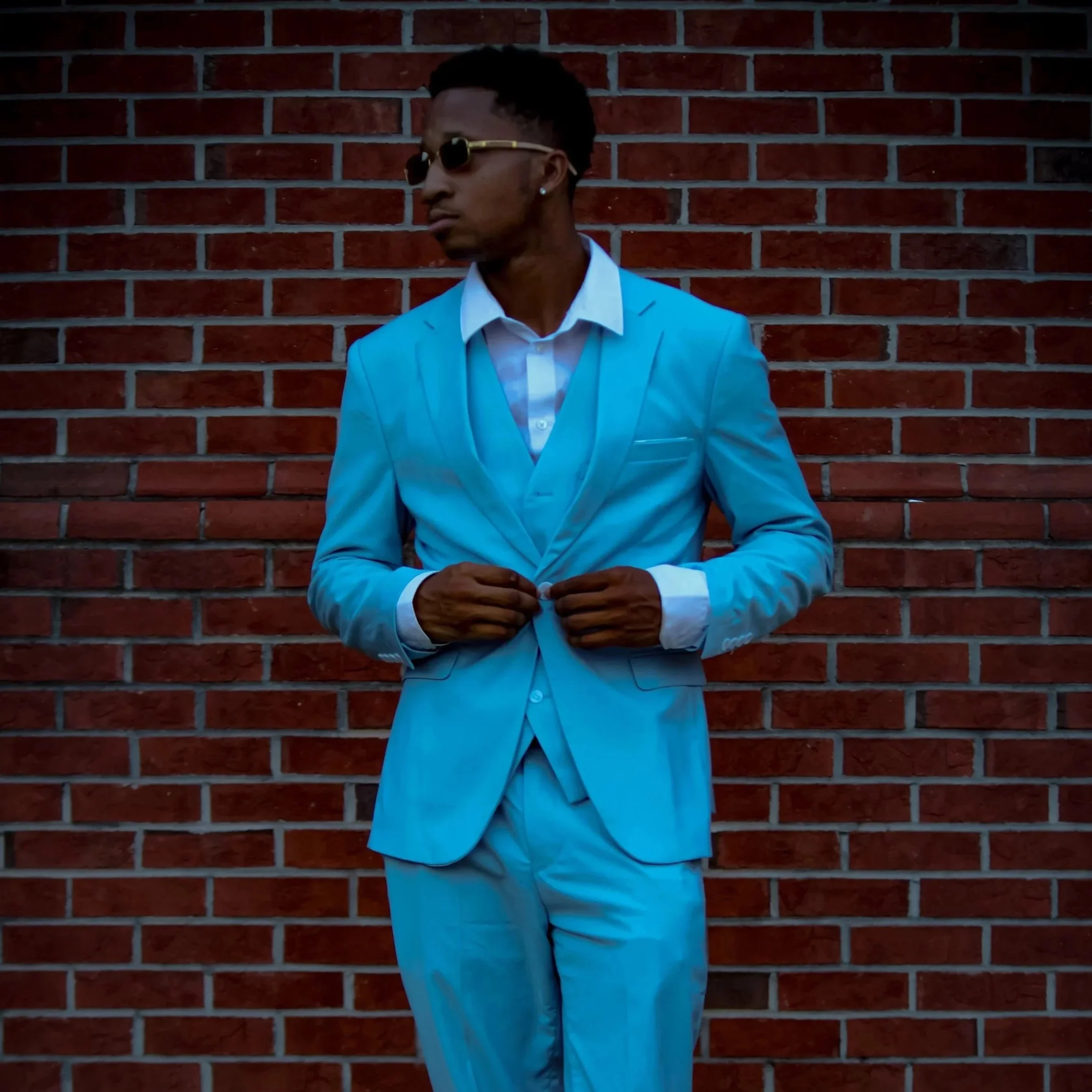 A man in a light blue suit with a white shirt, wearing sunglasses, stands against a brick wall, adjusting his suit jacket. Fayetteville, NC