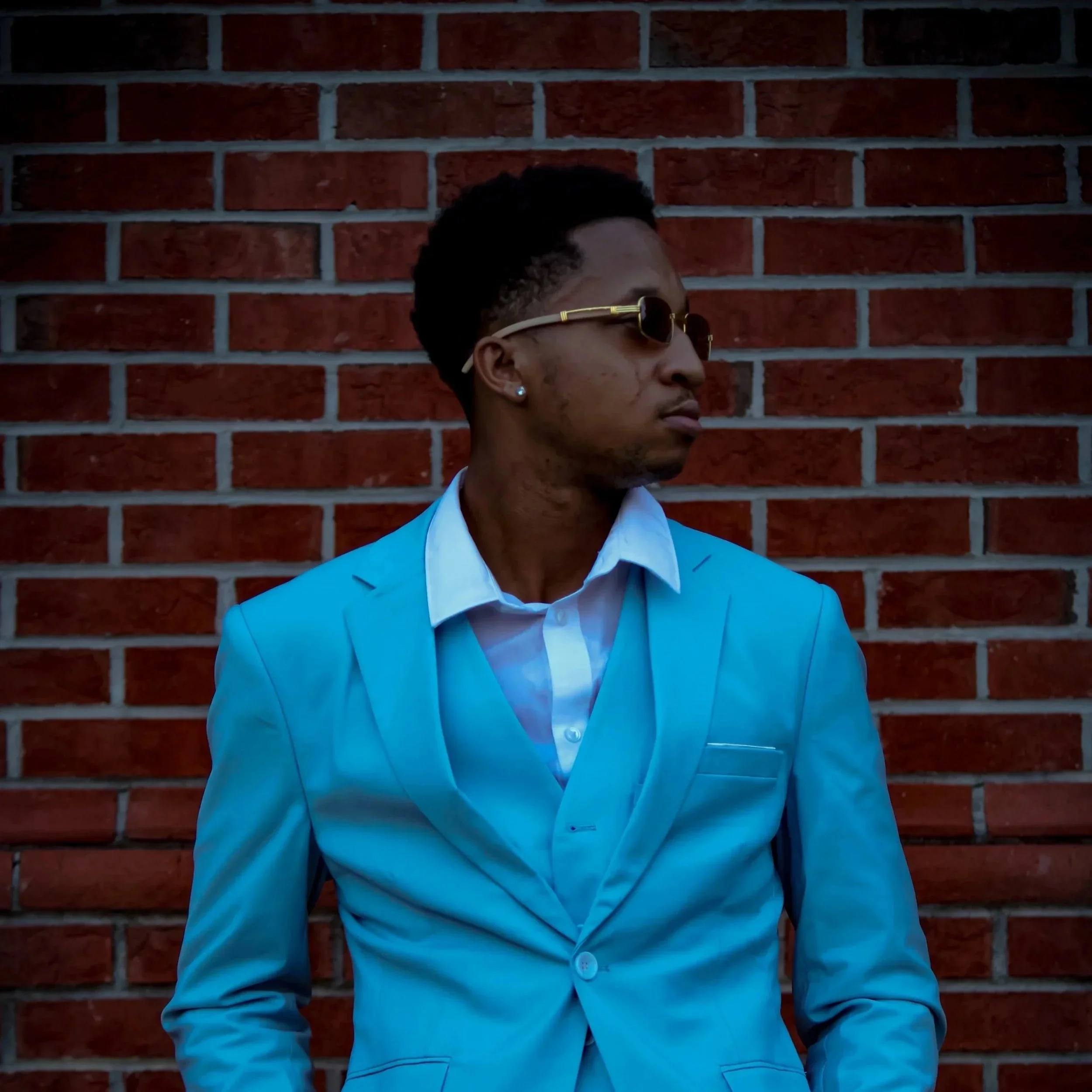 A young man with short, styled hair, sunglasses, and earrings wearing a light blue suit and white shirt stands against a red brick wall, looking to the side. Fayetteville, NC