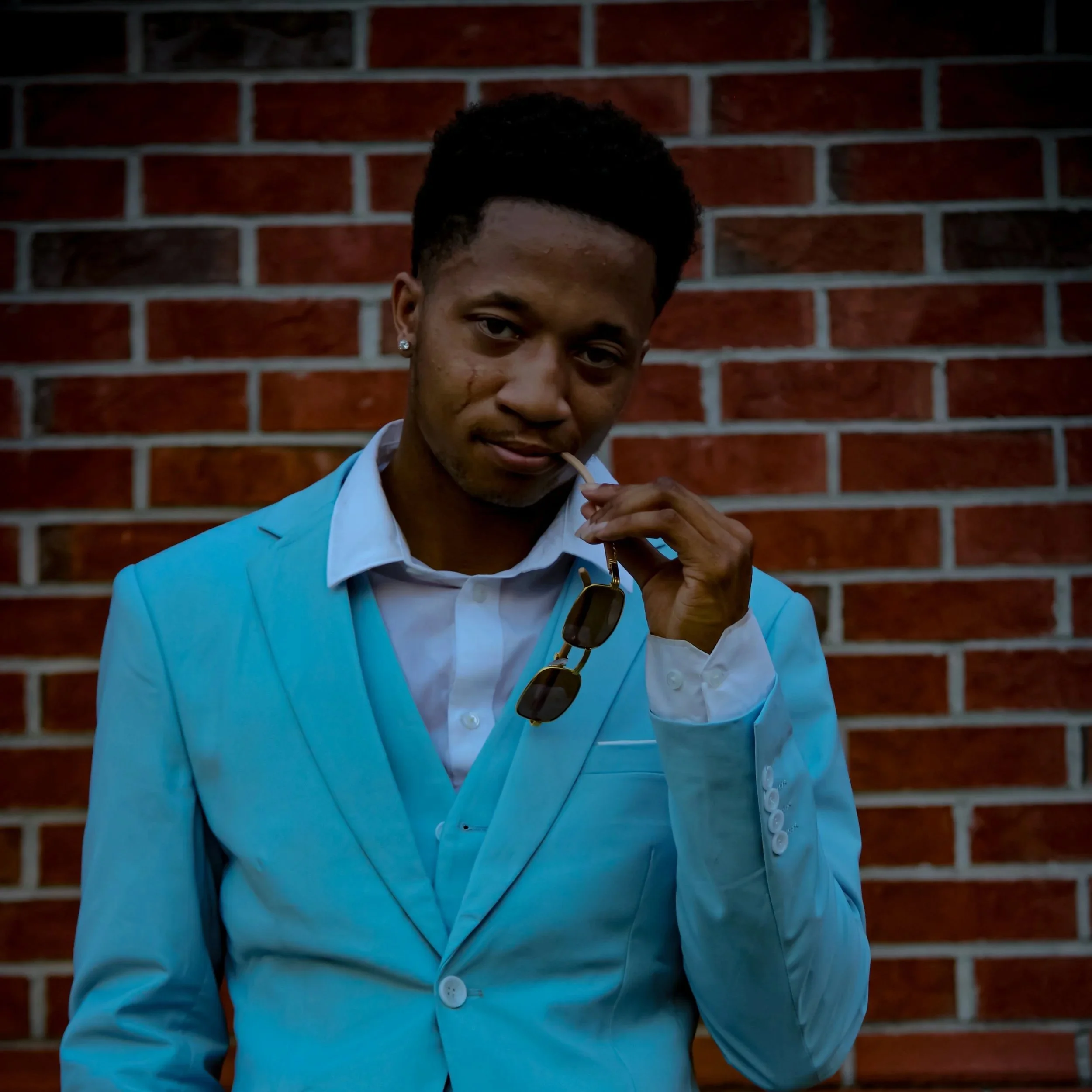 Young man in a light blue suit standing against a red brick wall, holding sunglasses in his left hand and a lollipop in his mouth, looking at the camera. Fayetteville, NC