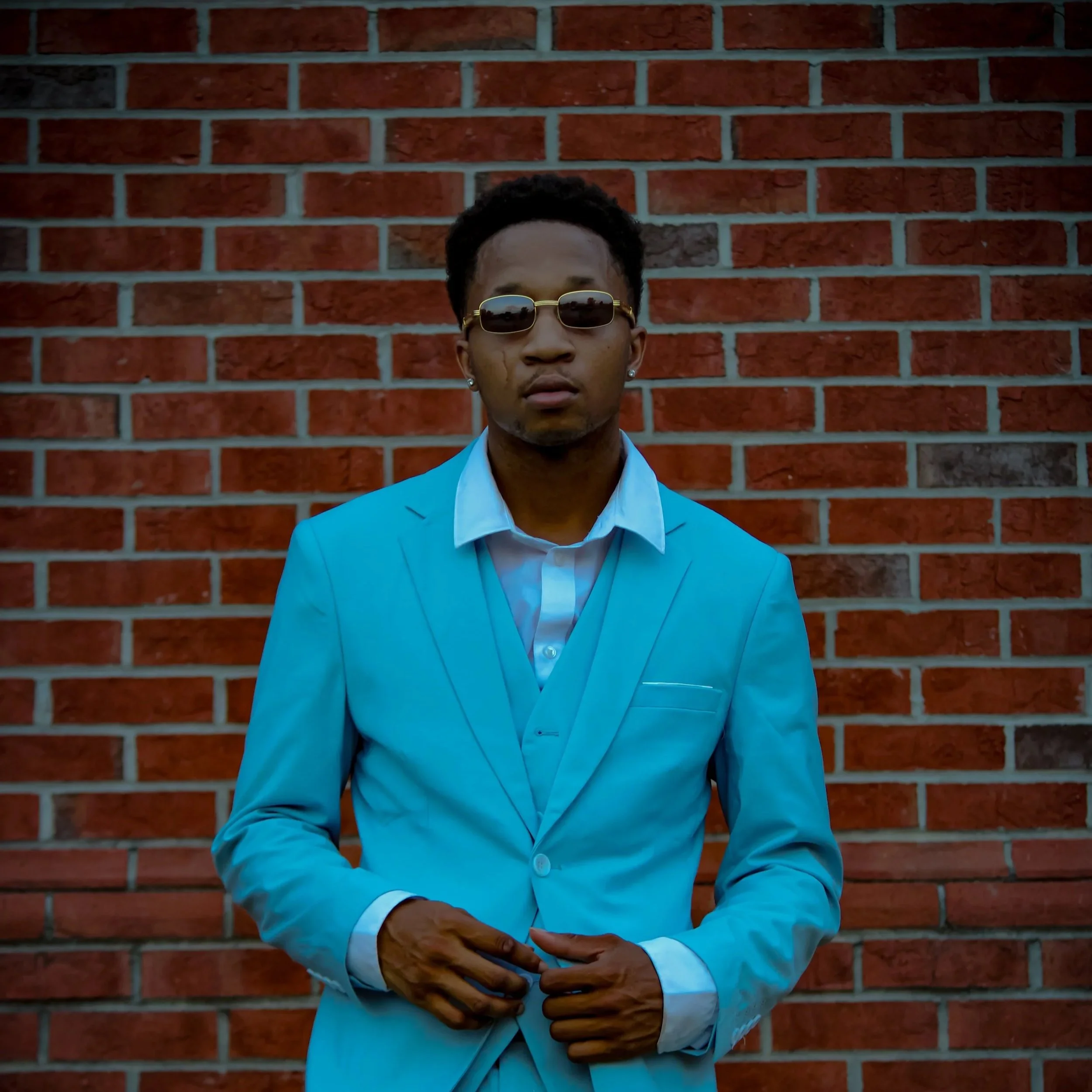 A young man in a light blue suit and white shirt Standing in front of a red brick wall wearing sunglasses. Fayetteville, NC