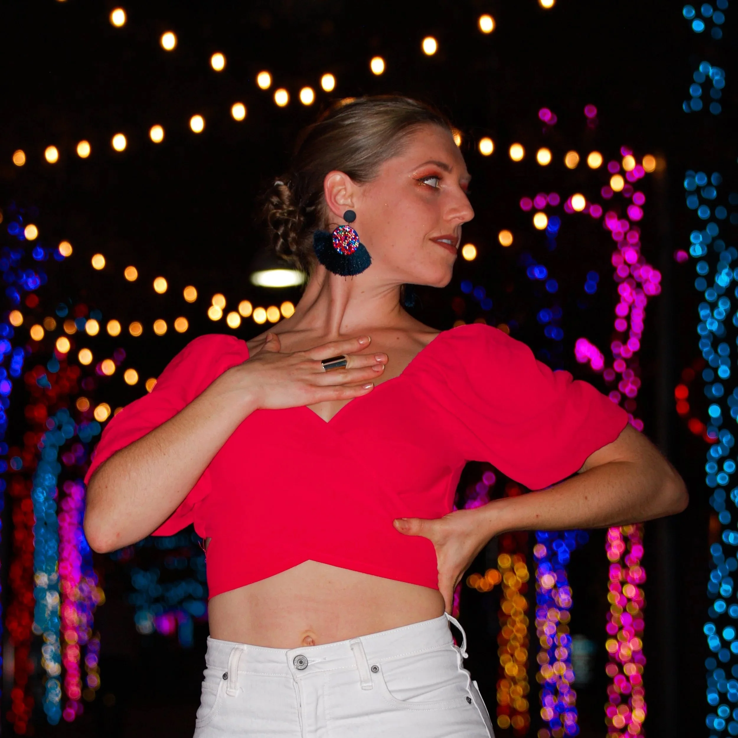 A woman in a red crop top with puffed sleeves and white pants, posing with one hand on her chest and the other on her hip, against a backdrop of colorful, blurred lights at night. Austin, TX