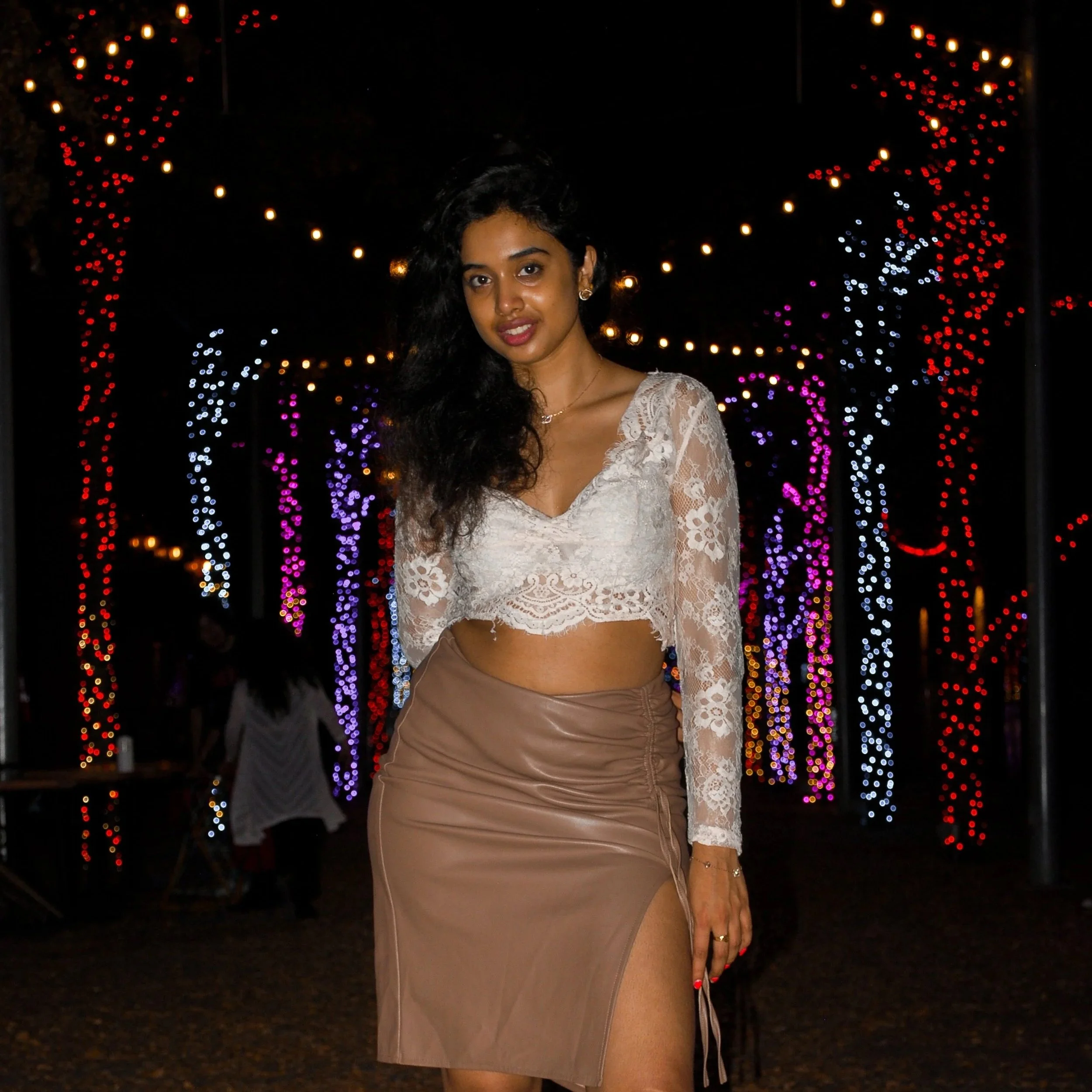 Young woman with long black hair wearing a lace crop top and a beige leather skirt with a slit, standing in front of colorful string lights at night. Austin, TX