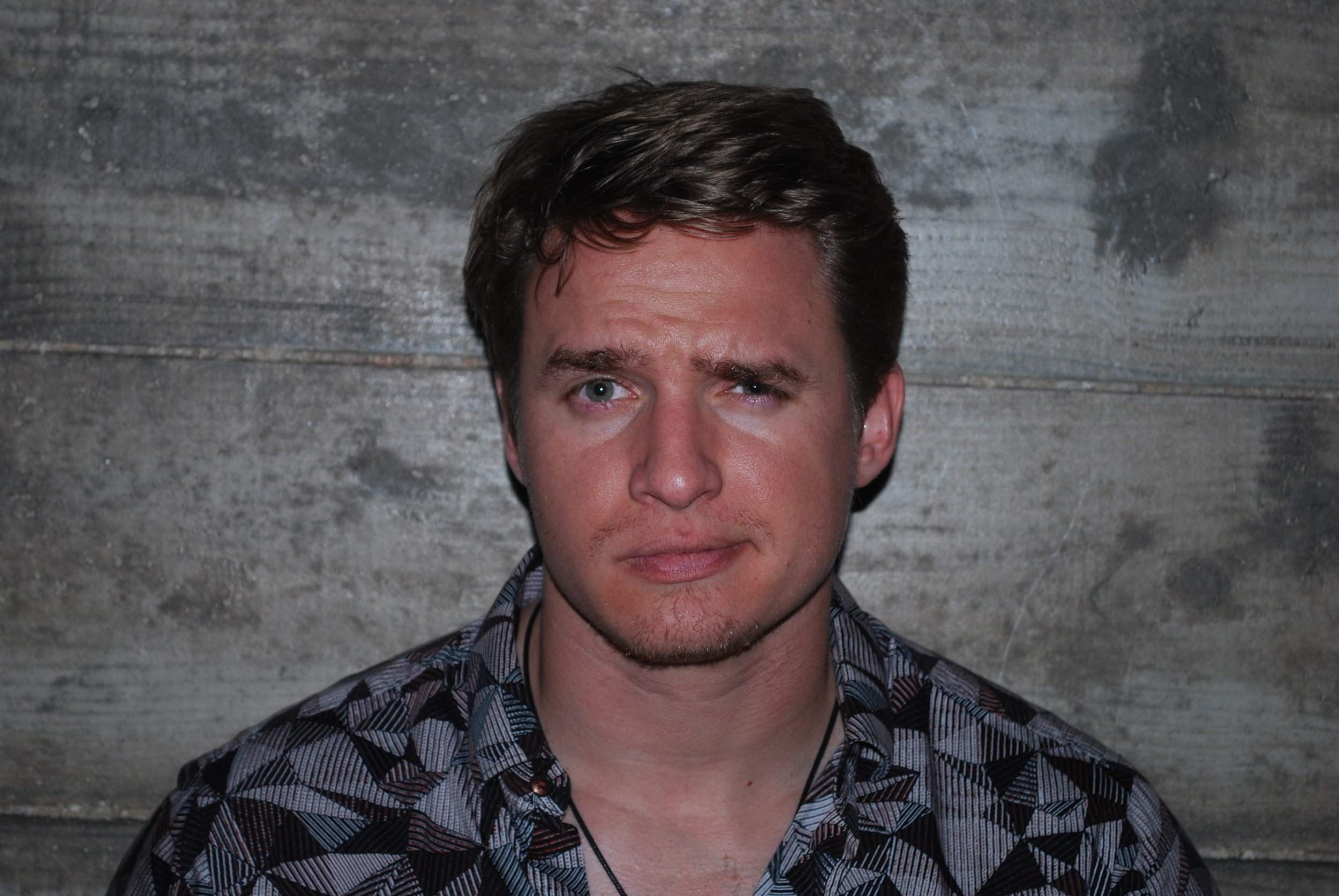 A man with light skin, brown hair, and a slight beard winking and making a skeptical facial expression in front of a wooden background. Austin, TX