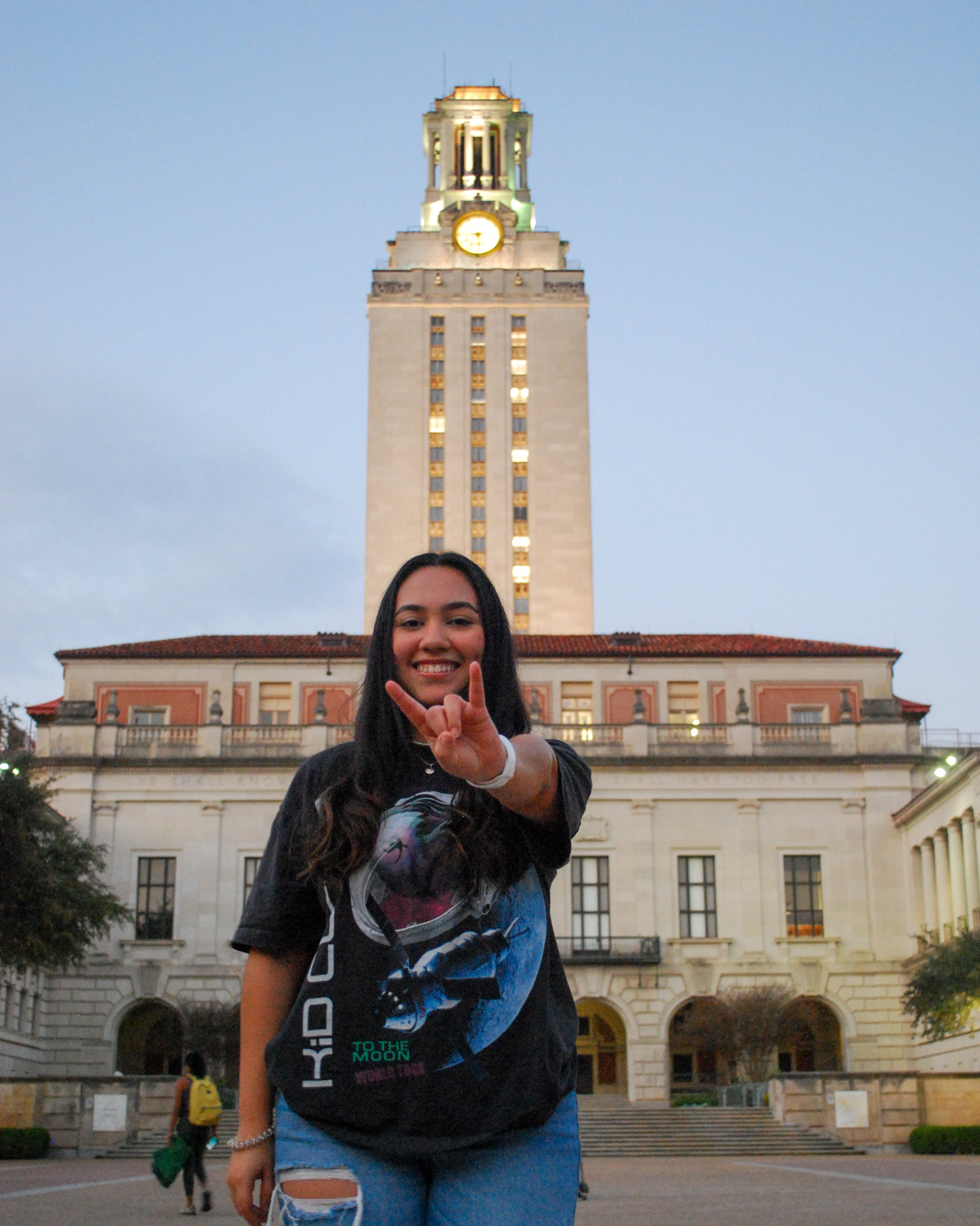 A young woman smiling and making a gesture with her hand in front of the University of California, Berkeley campus with the Bell Tower at dusk. Austin, TX