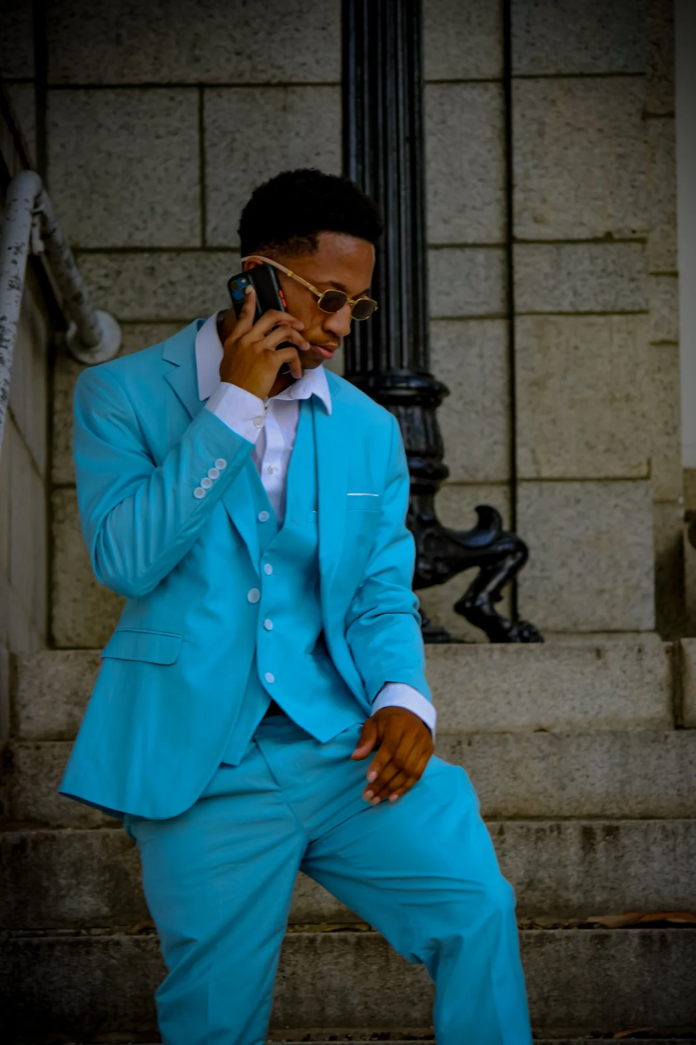 A man in a bright blue suit and sunglasses is talking on a cellphone while posing on stone stairs outside a building. Fayetteville, NC