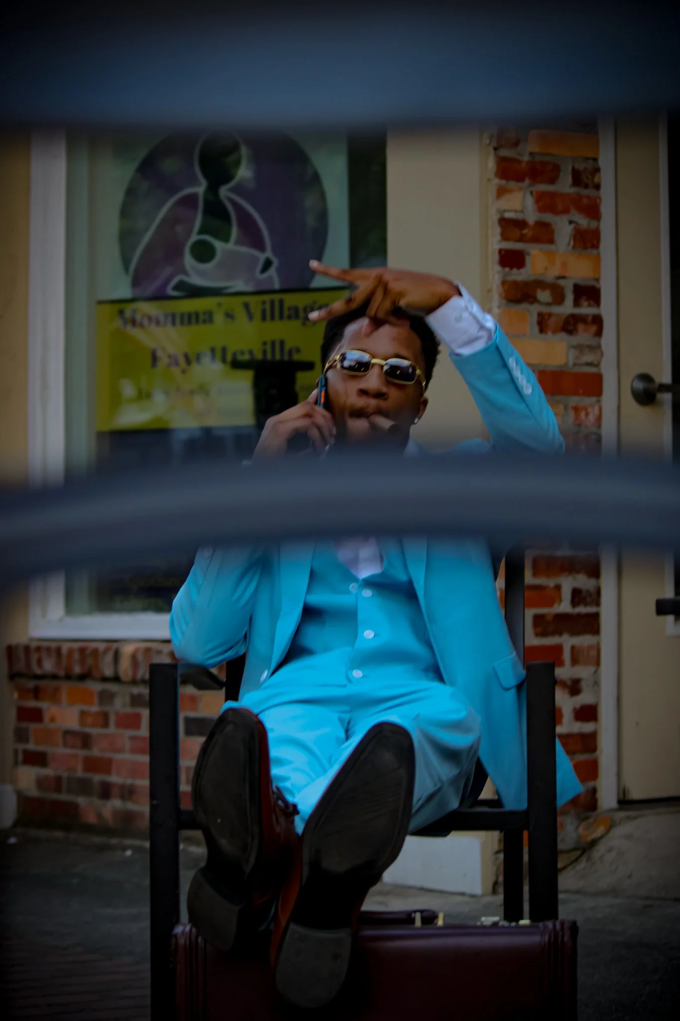 A young man in a light blue suit and sunglasses is sitting outside, talking on a cellphone, with his legs up on a suitcase. He is taking a selfie or looking through a gap in a fence, with a building and a sign in the background. Fayetteville, NC