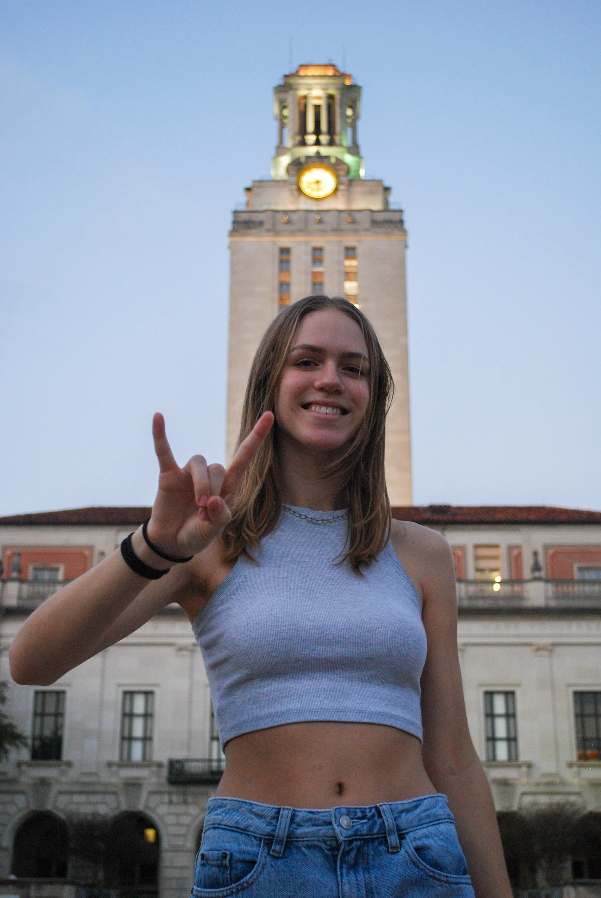 Young woman smiling and making the rock on hand gesture in front of a tall clock tower during dusk. Austin, TX
