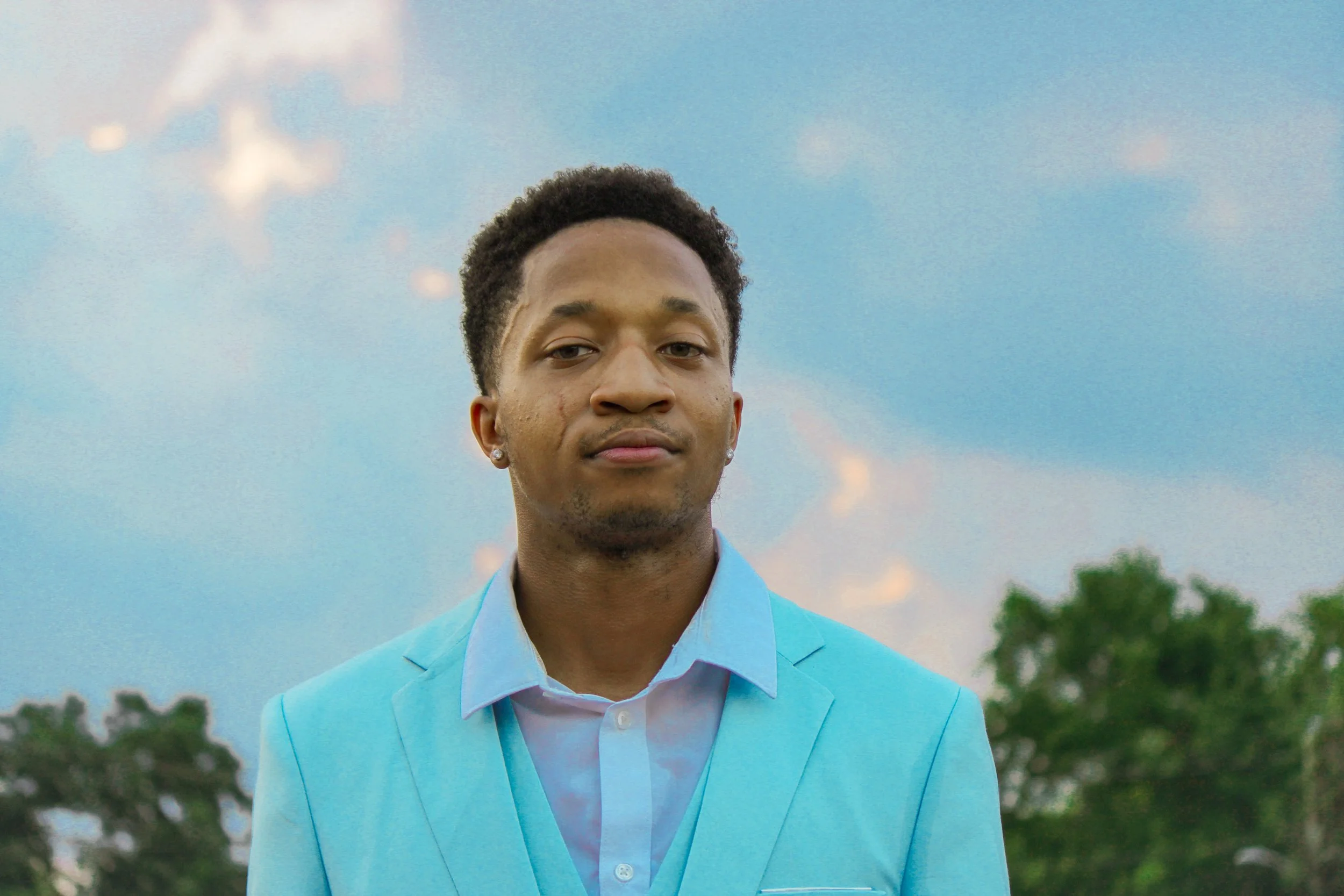 A young man with short curly hair, wearing a light blue suit and white shirt, standing outdoors with a background of a cloudy sky and trees. Fayetteville, NC
