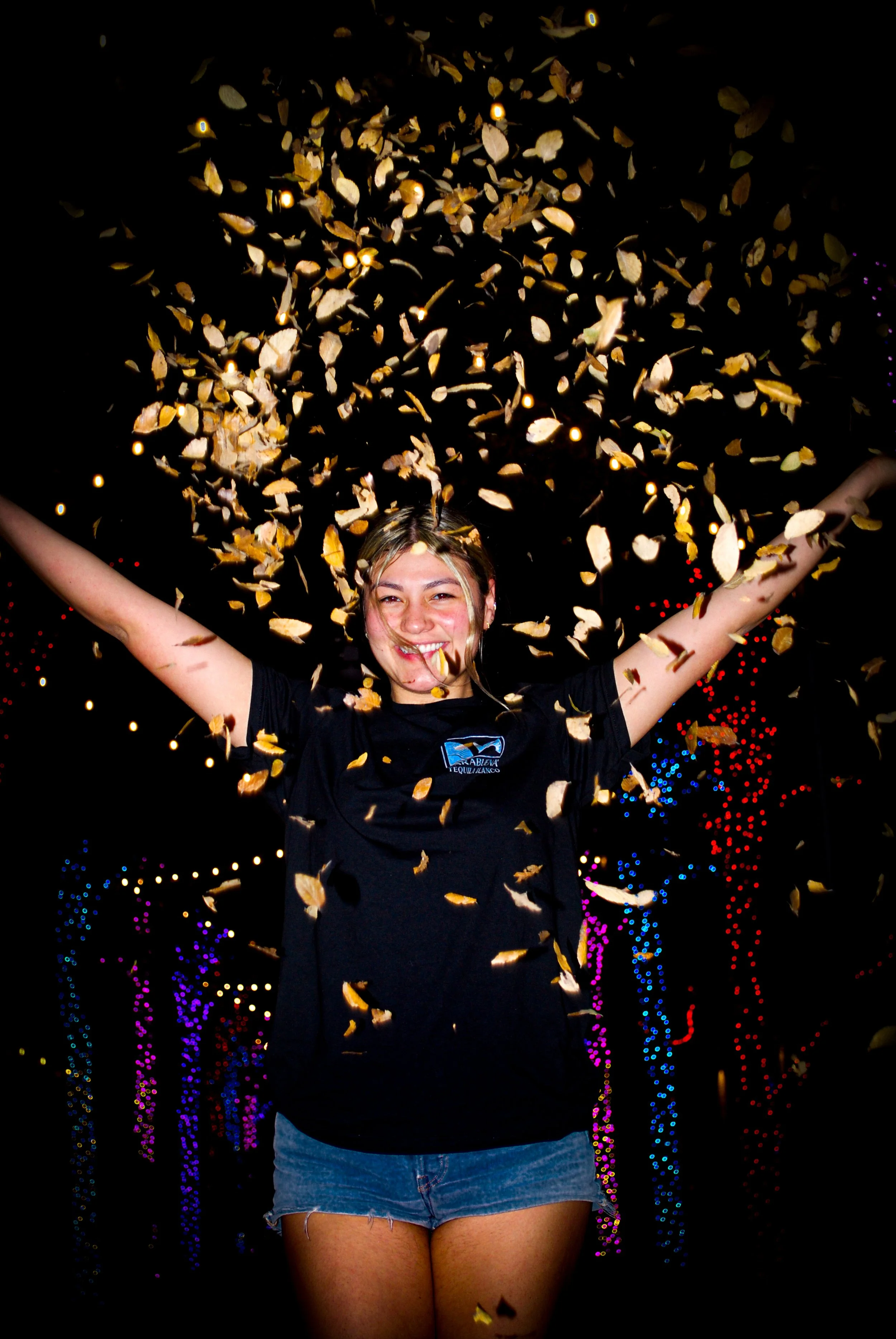 A woman smiling with arms raised as confetti falls around her at night, with colorful lights in the background. Austin, TX