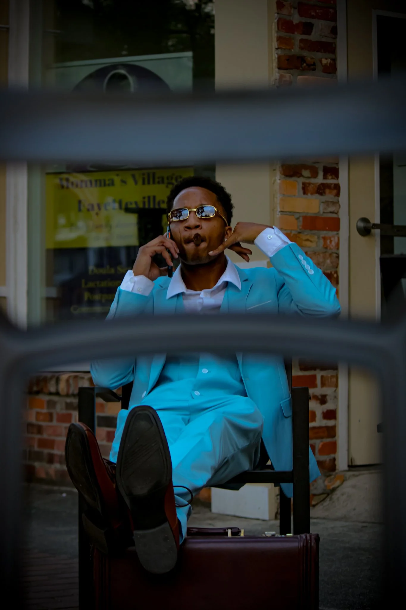 A man in a bright blue suit and sunglasses talking on a cellphone, sitting on a chair outside a building, distinguished by brick wall details, with a yellow sign behind him. The photo is taken through metal bars. Fayetteville, NC