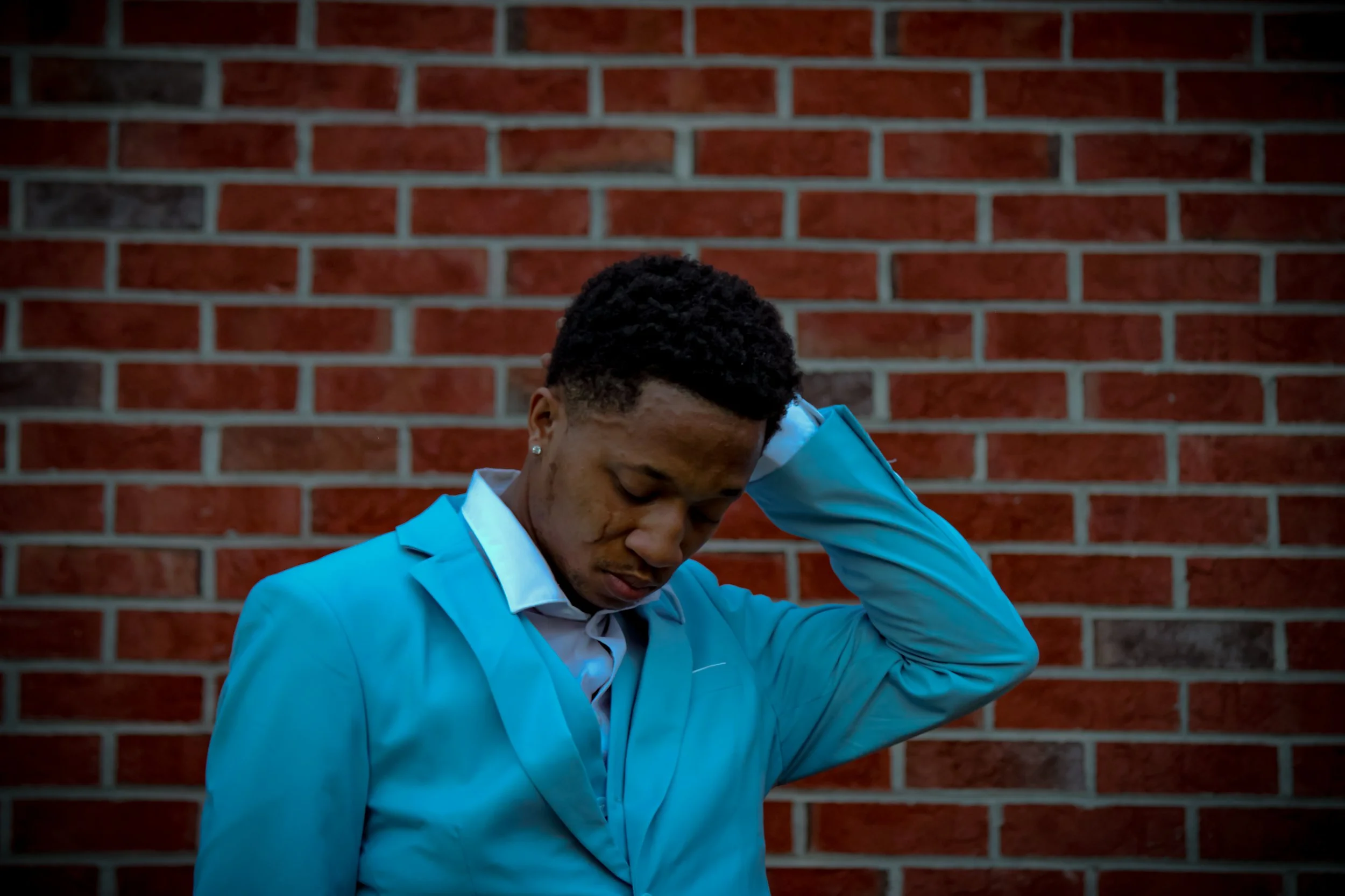 Young man wearing a light blue suit and white shirt standing against a brick wall, looking down with a contemplative expression. 
Fayetteville, NC