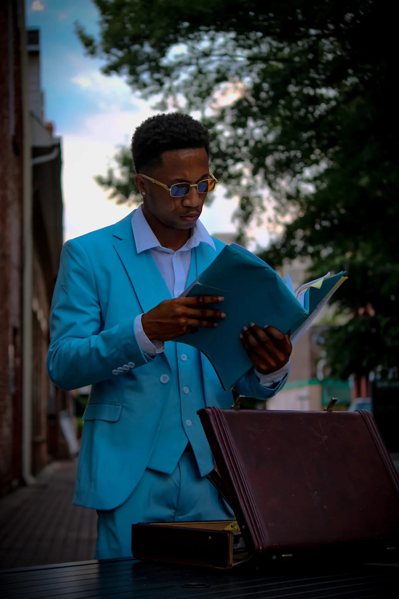 A young man in a light blue suit and sunglasses reading a folder during daytime outdoors. Fayetteville, NC