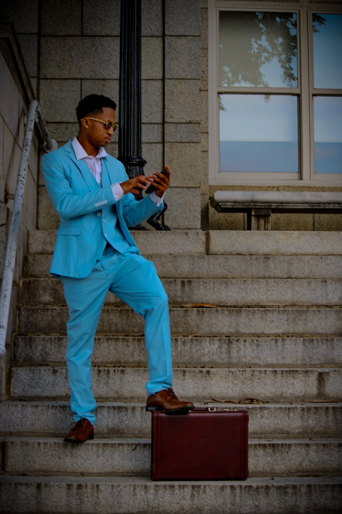 A man in a light blue suit stands on steps outside a building, using a smartphone. A brown suitcase is positioned on the step in front of him. The scene appears to be near a waterfront, as the window reflects the ocean. Fayetteville, NC