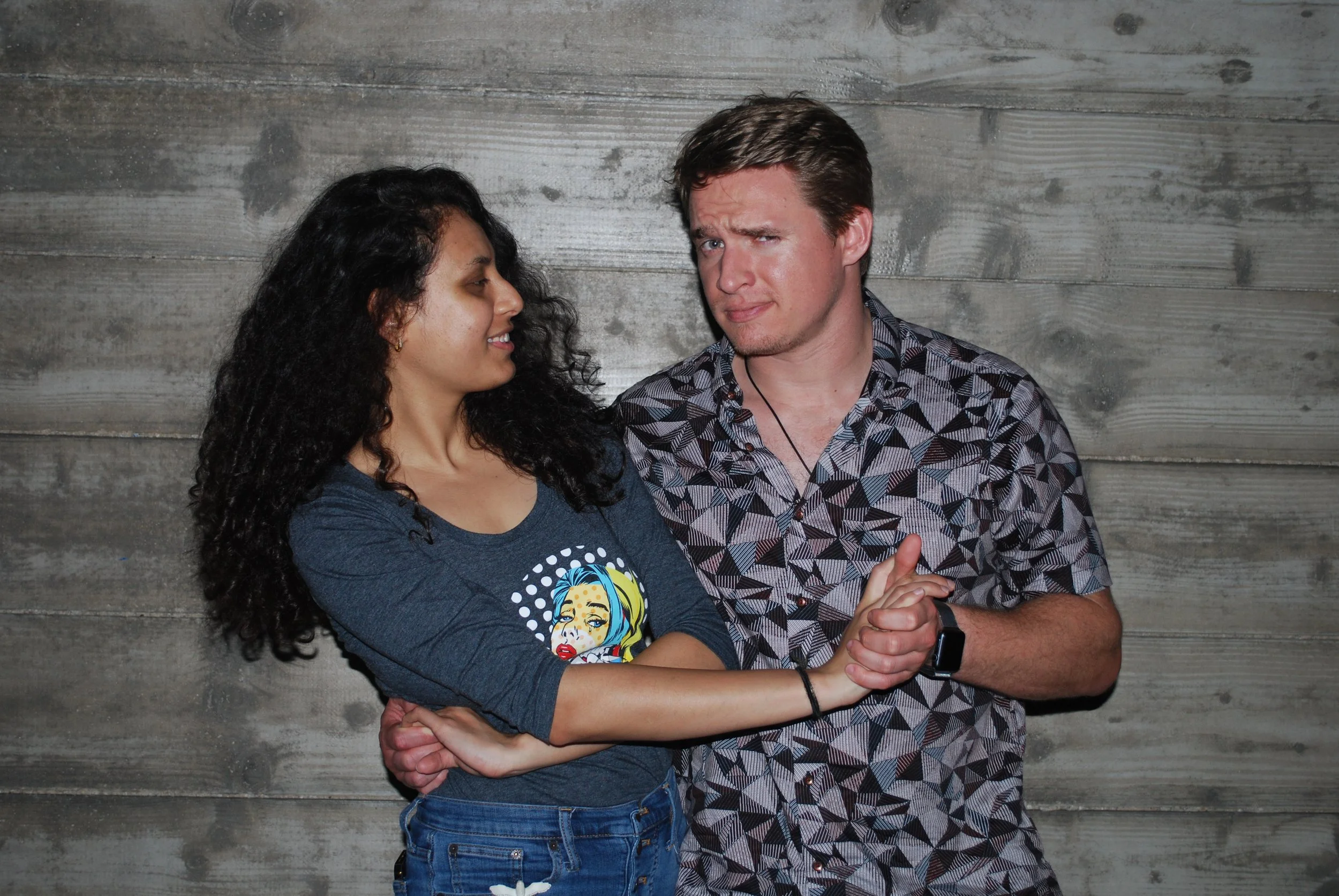 A woman and a man are standing close together against a wooden wall. The woman has curly black hair, is wearing a gray shirt with a colorful graphic, and blue jeans, smiling with her arms crossed. The man has short brown hair, is wearing a patterned 