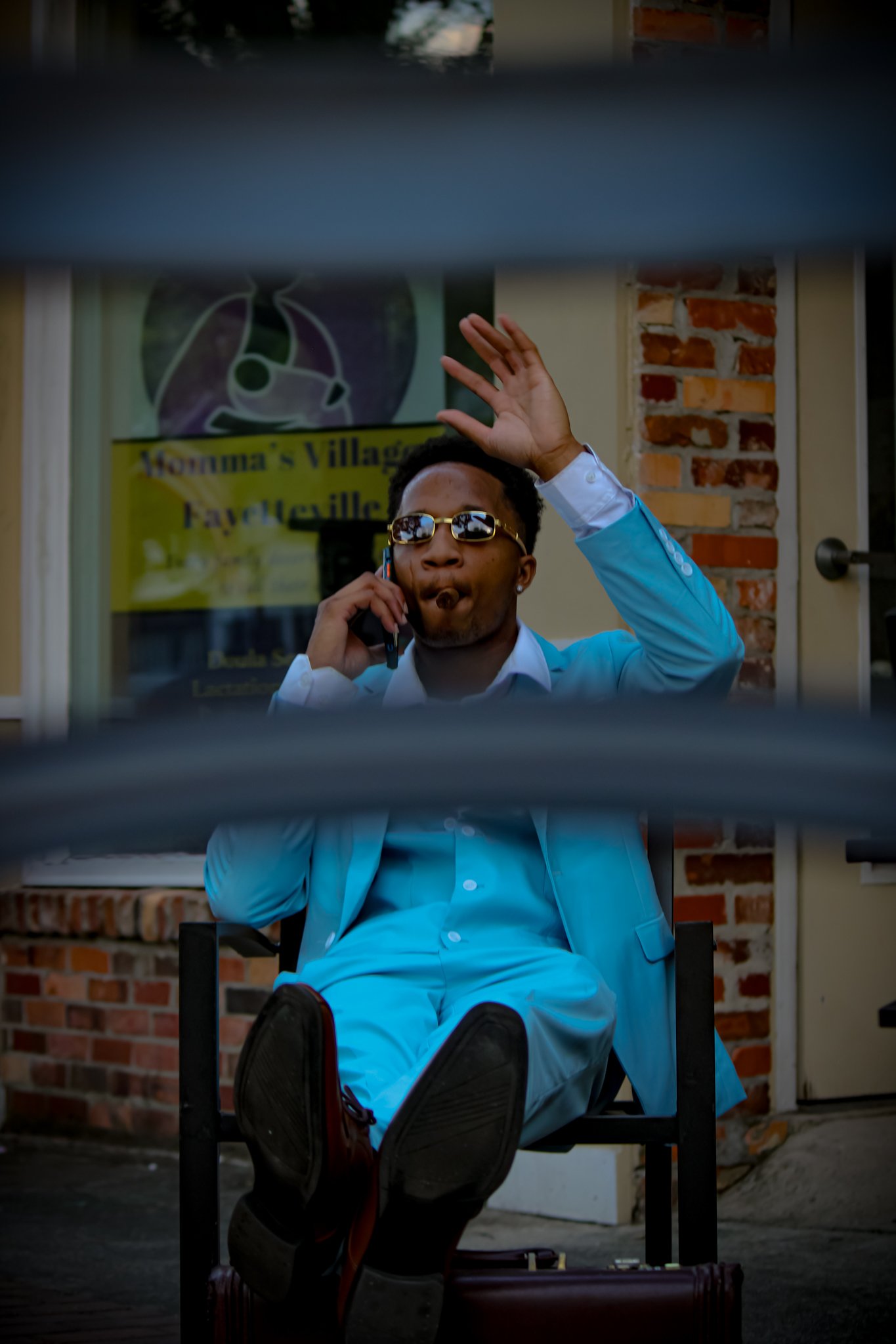 A man with sunglasses and a white suit, sitting outside, talking on a cellphone, raising one hand, seen through a window or gap. 
Fayetteville, NC