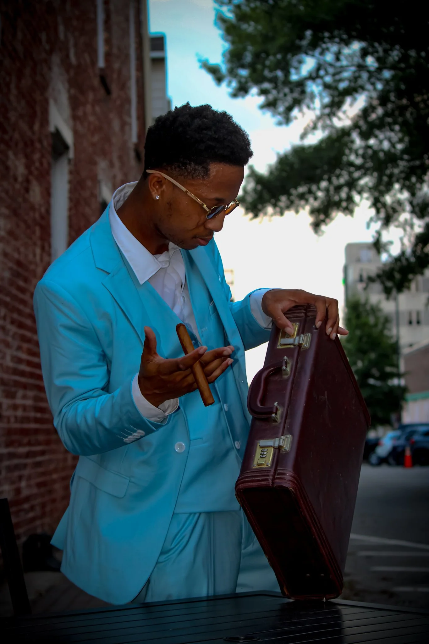 A man dressed in a light blue suit, holding a cigar and opening a vintage briefcase outdoors, with a brick wall on one side and trees in the background. Fayetteville, NC
