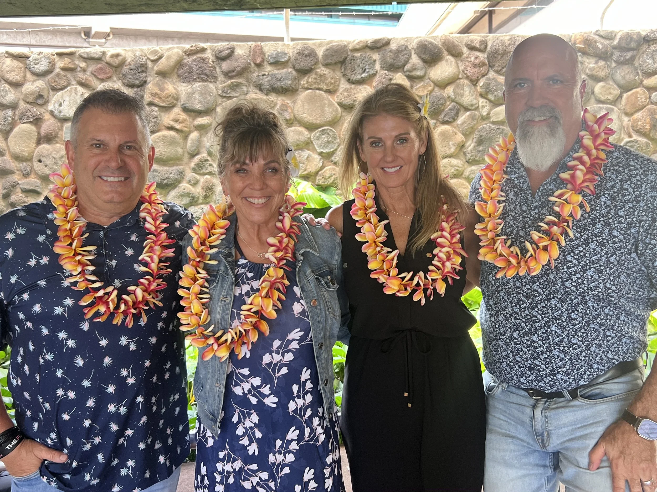 Four adults standing together, all wearing colorful lei necklaces, smiling in front of a stone wall with tropical plants.