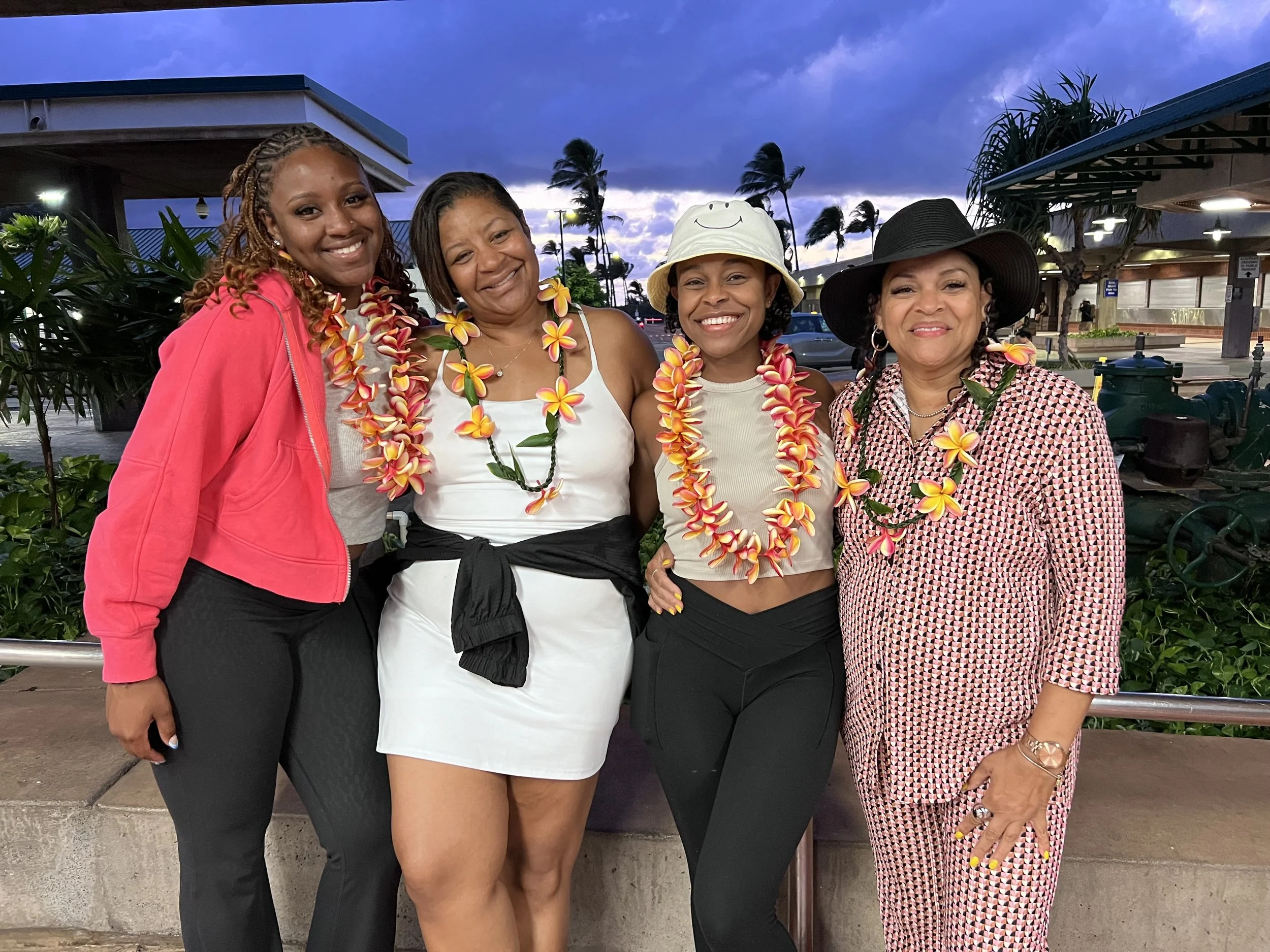 Four women standing outdoors at sunset or dusk, wearing colorful leis, and dressed in casual summer attire, with palm trees and a building in the background.