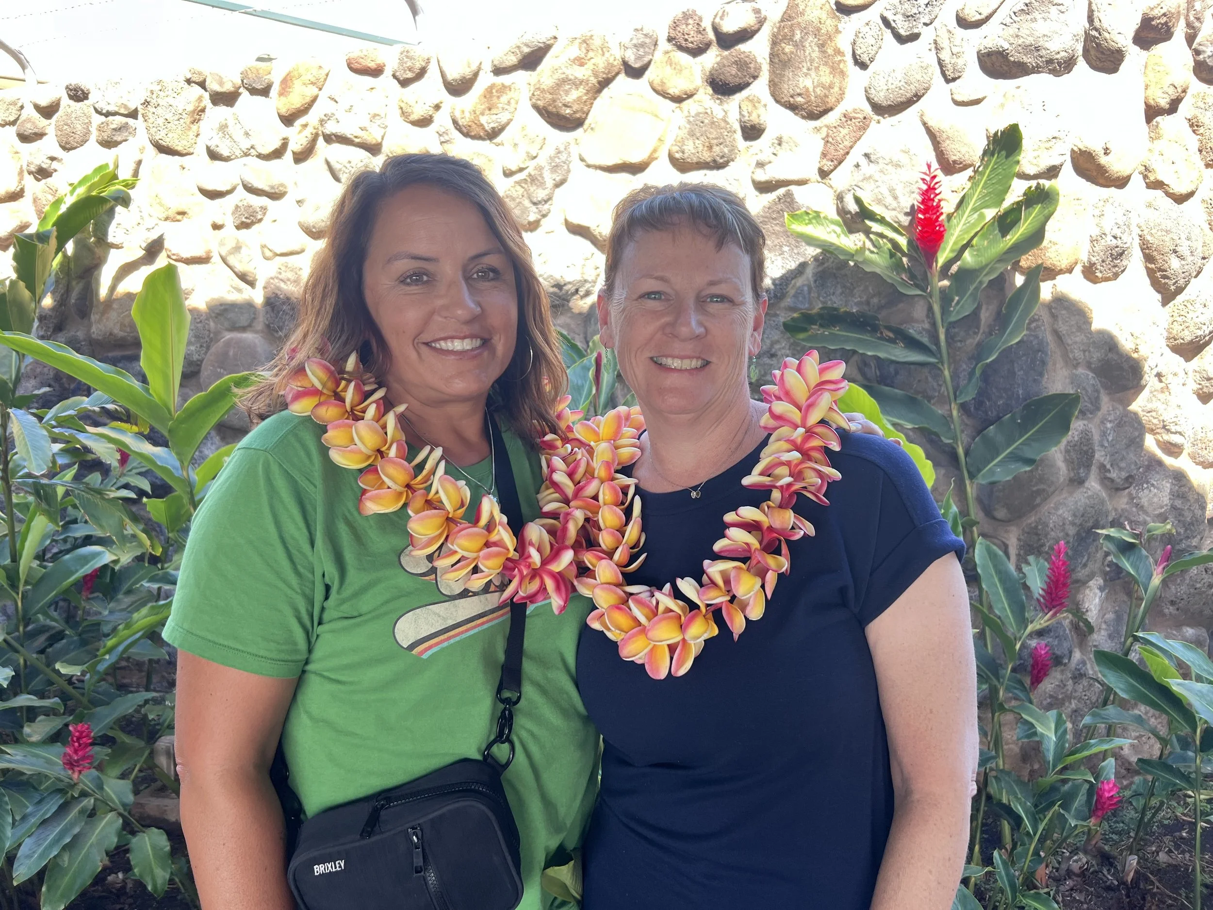 Two women smiling, wearing flower leis, standing in front of a stone wall with tropical plants, one in a green t-shirt and the other in a navy t-shirt.