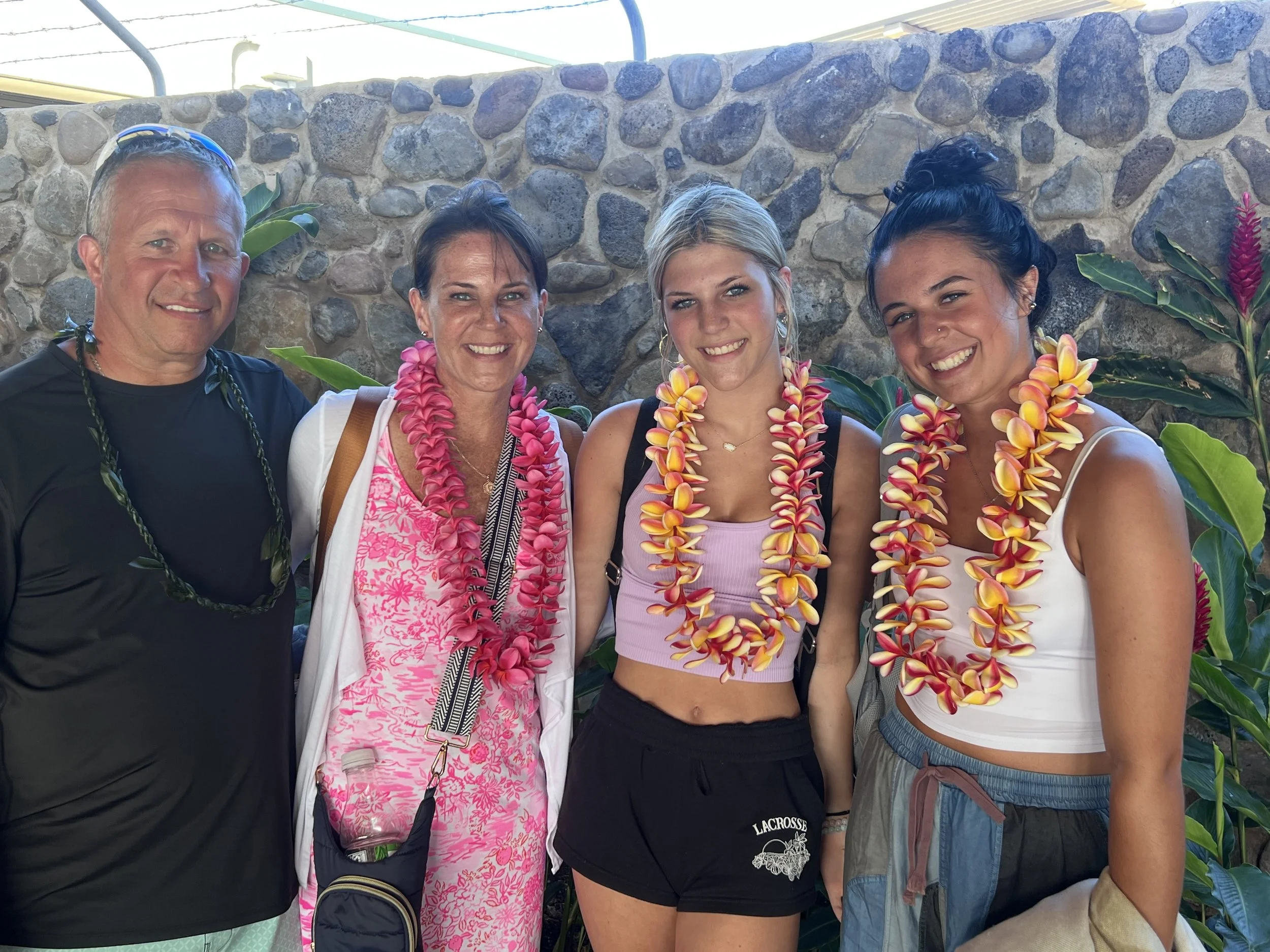 Four people standing outdoors against a stone wall, all wearing floral leis. They are smiling and appear to be enjoying a tropical-themed celebration.
