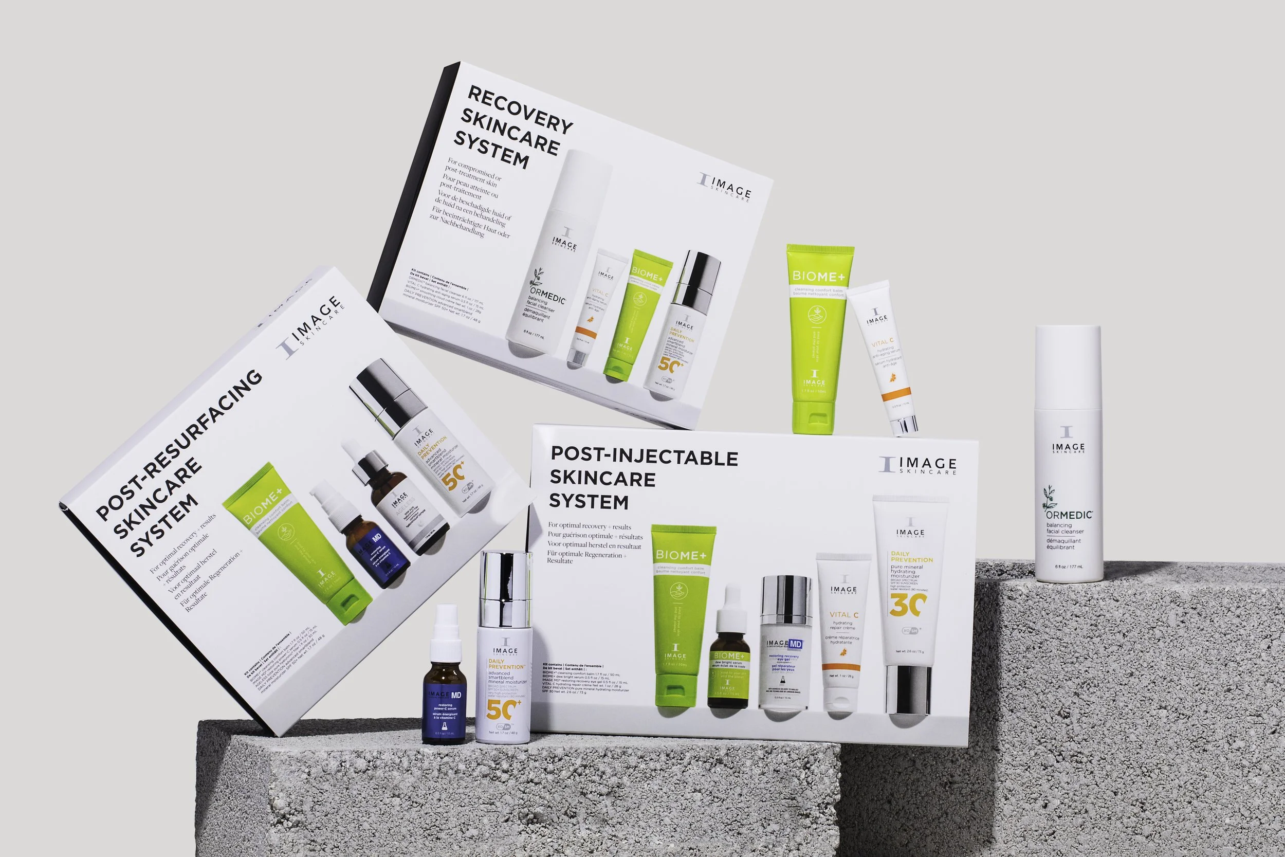 Skincare products displayed on gray concrete blocks with white background, including boxes and bottles labeled with skincare system names like Post-Resurfacing, Post-Injectable, and Recovery System, along with individual products in tubes and bottles.
