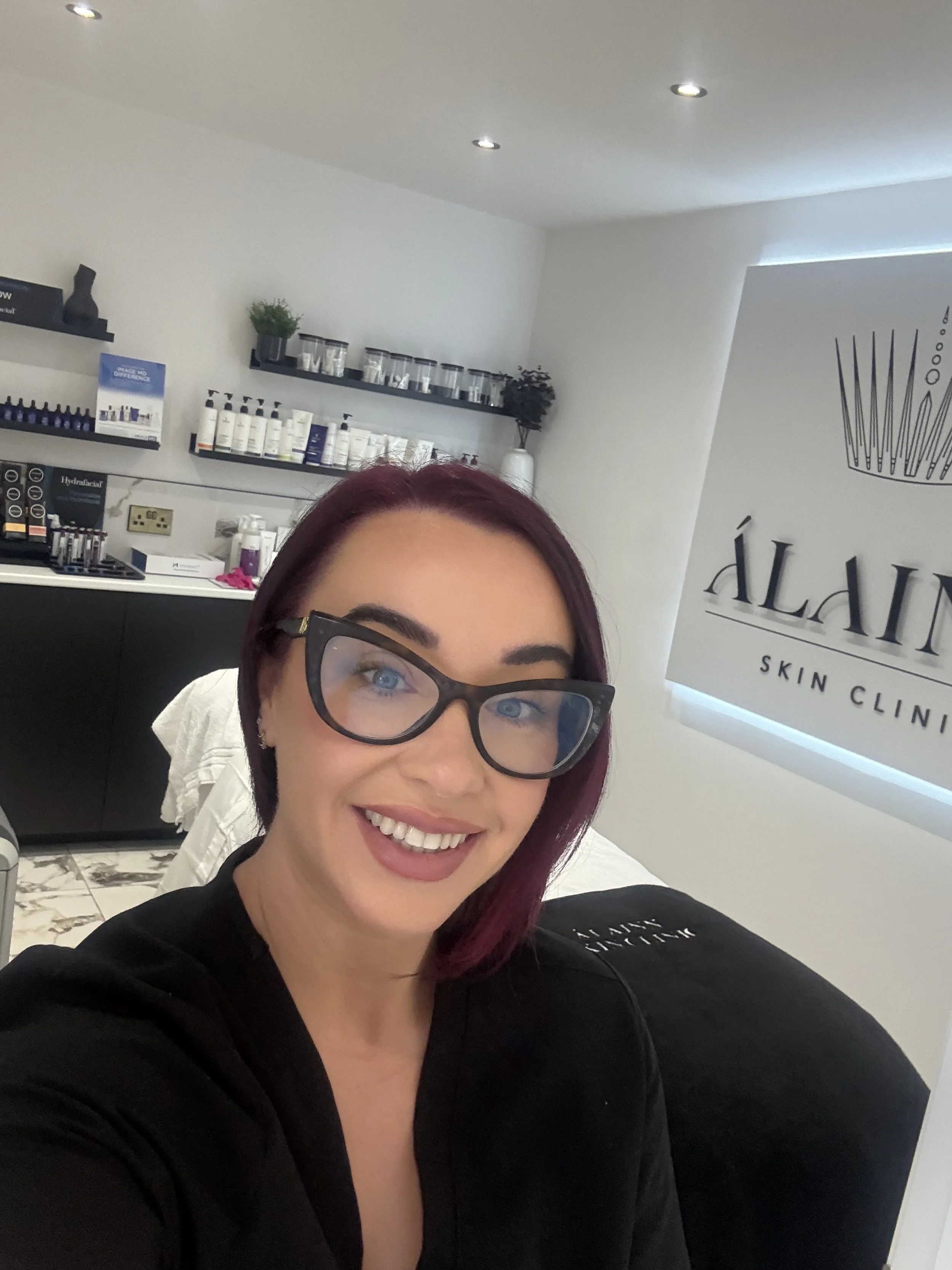 A woman with purple hair and glasses smiling in a skin clinic room with skincare products on shelves and a logo on the wall.