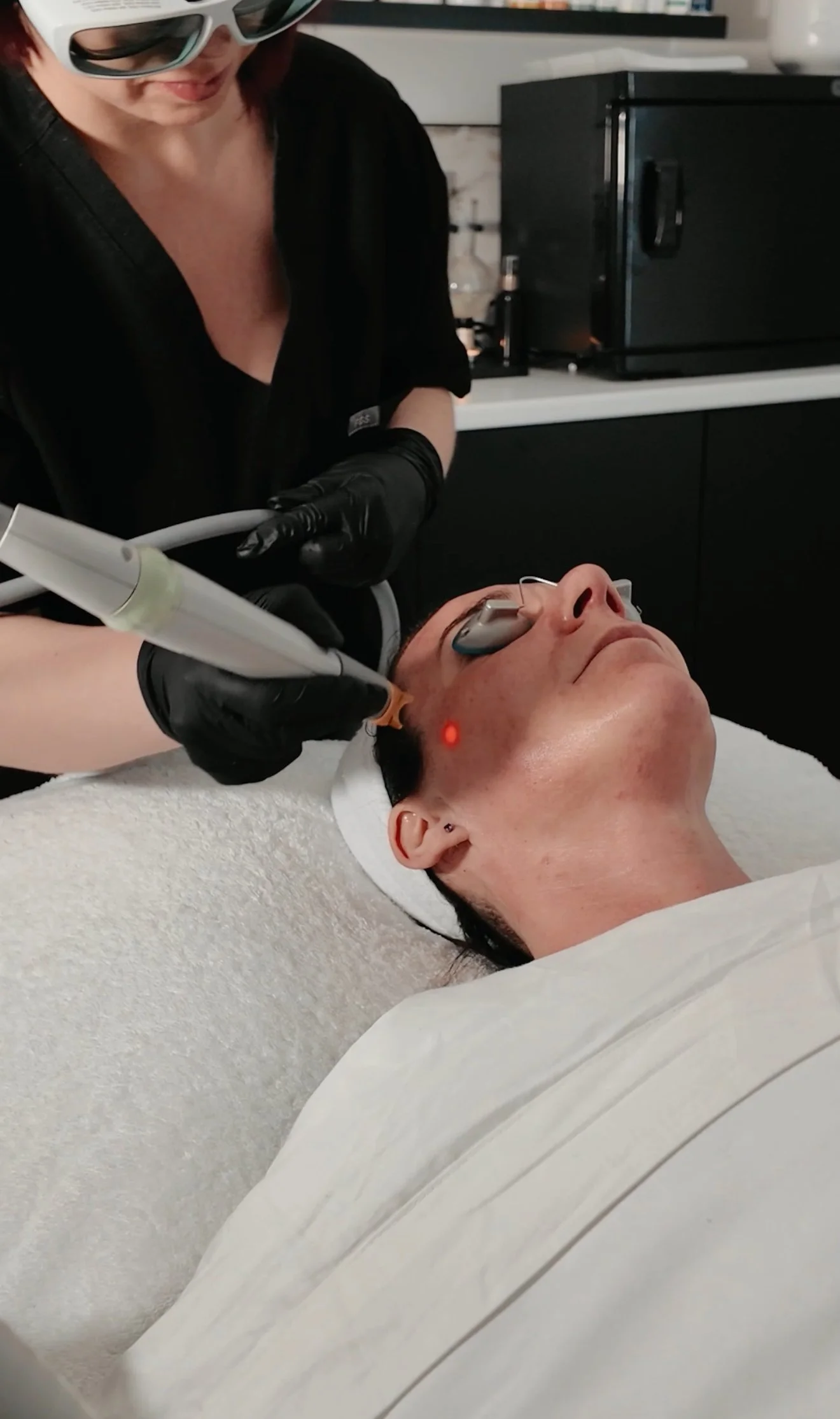 A person receiving a laser skin treatment on their face while lying on a treatment bed, with a technician performing the procedure with a handheld device, wearing black gloves and protective eyewear.