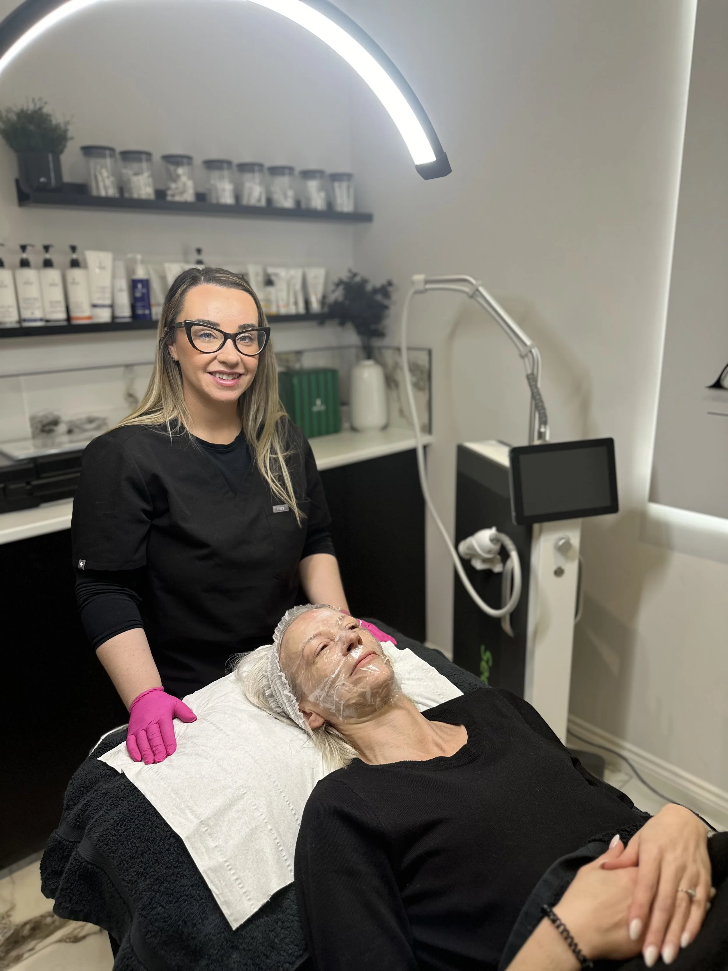 A woman lying on a treatment bed with a facial mask, receiving a facial treatment from a skincare professional in a spa or clinic setting.