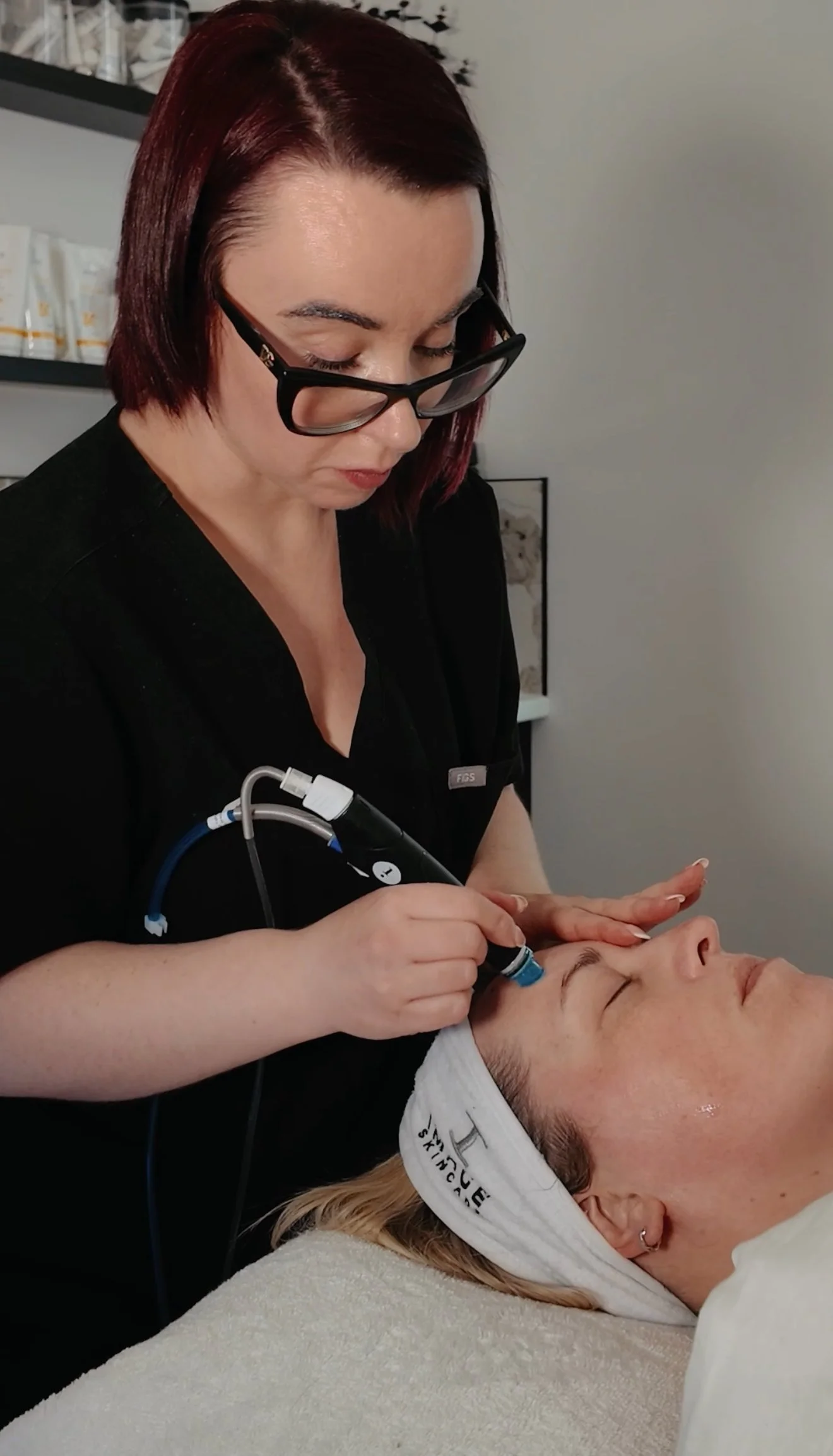 A woman receiving a facial treatment with a blue device while lying down with a headband, in a professional spa or clinic setting.