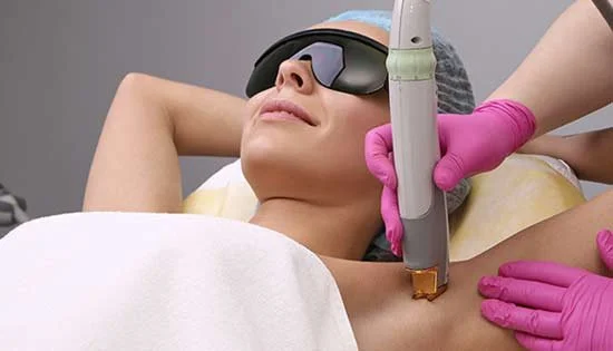 A woman receiving a laser treatment on her chest, wearing protective glasses, with a technician wearing pink gloves holding a laser device nearby.