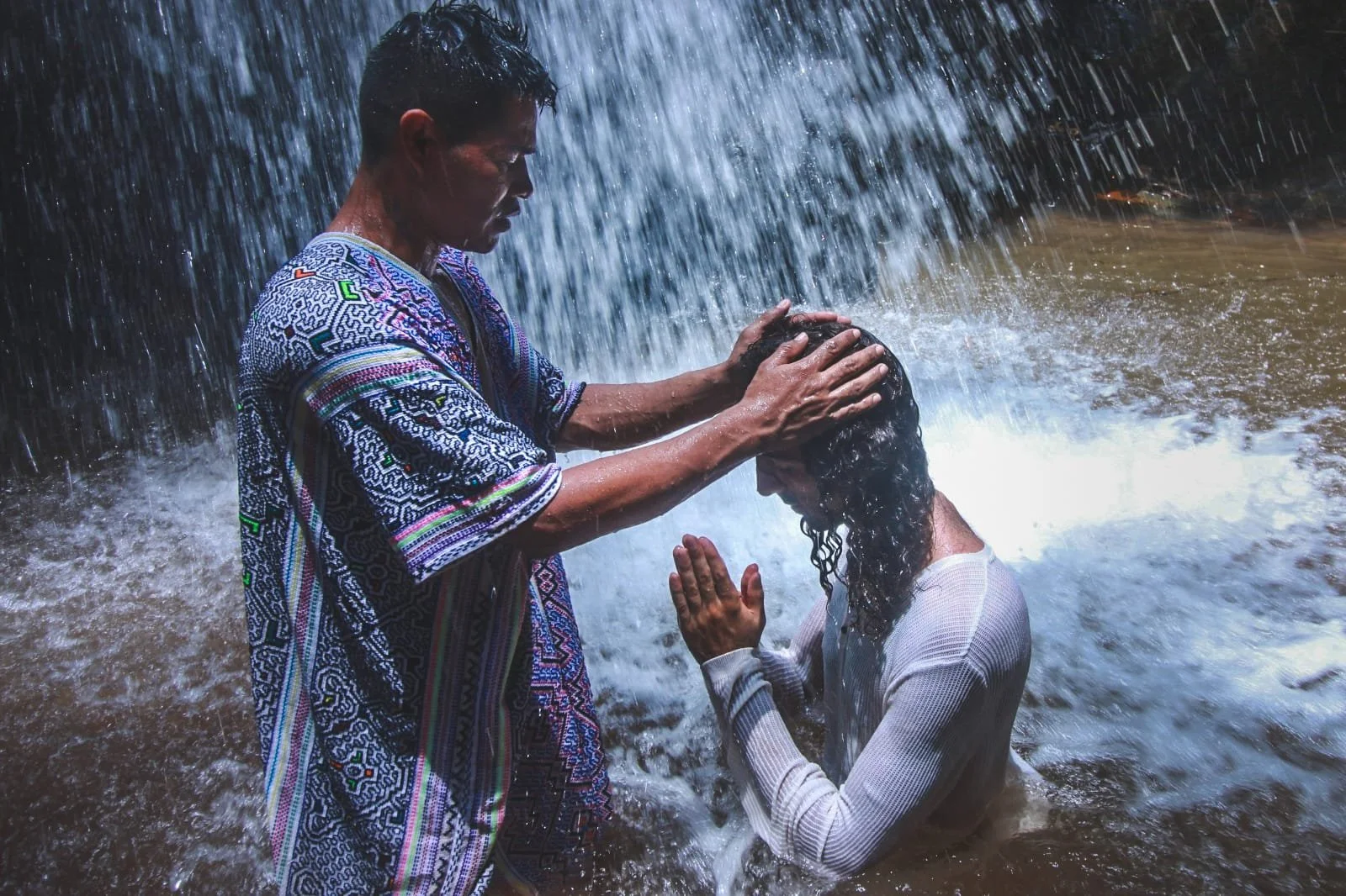Zwei Personen stehen unter einem Wasserfall, wobei eine Person die andere zu segnen oder zu taufen scheint. Die bekleidete Person kniet mit gefalteten Händen im Wasser, während die andere eine bunte Kleidung trägt und die Hände auf den Kopf des Knienden legt.