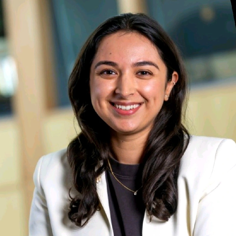 A woman smiling with long dark hair, wearing a white blazer and a black top, indoors with blurred background.