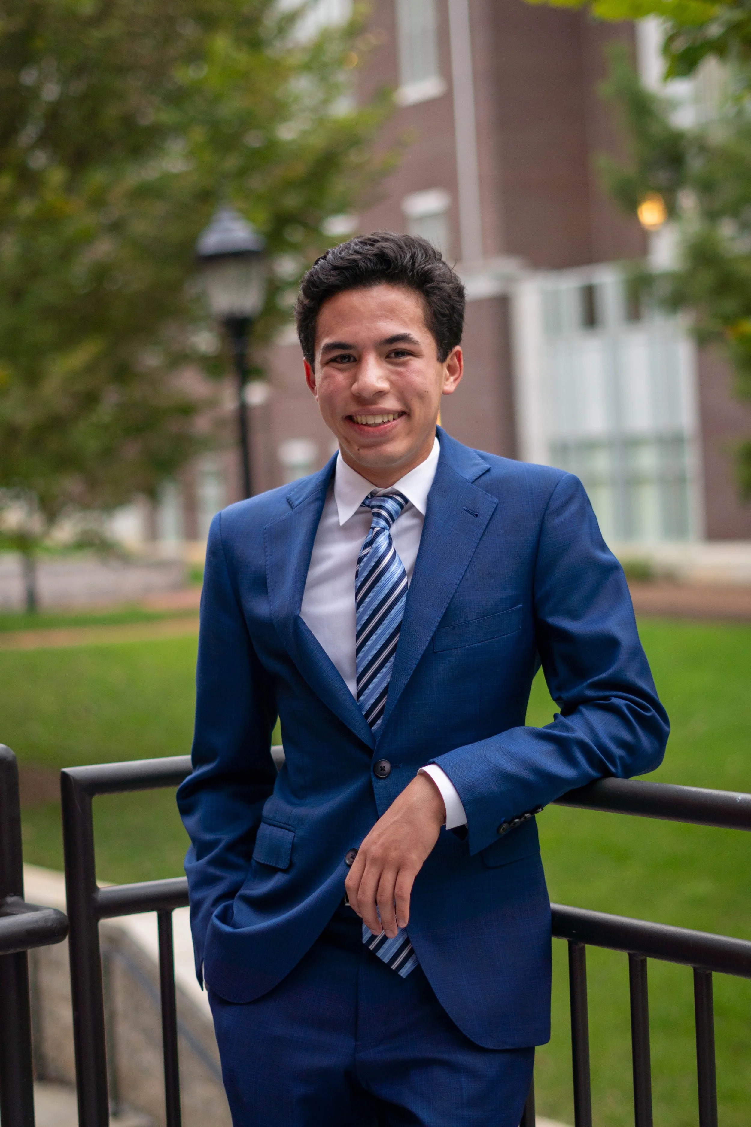 A young man in a blue suit, white shirt, and striped tie, standing outdoors near a black railing, smiling at the camera, with trees and buildings in the background.
