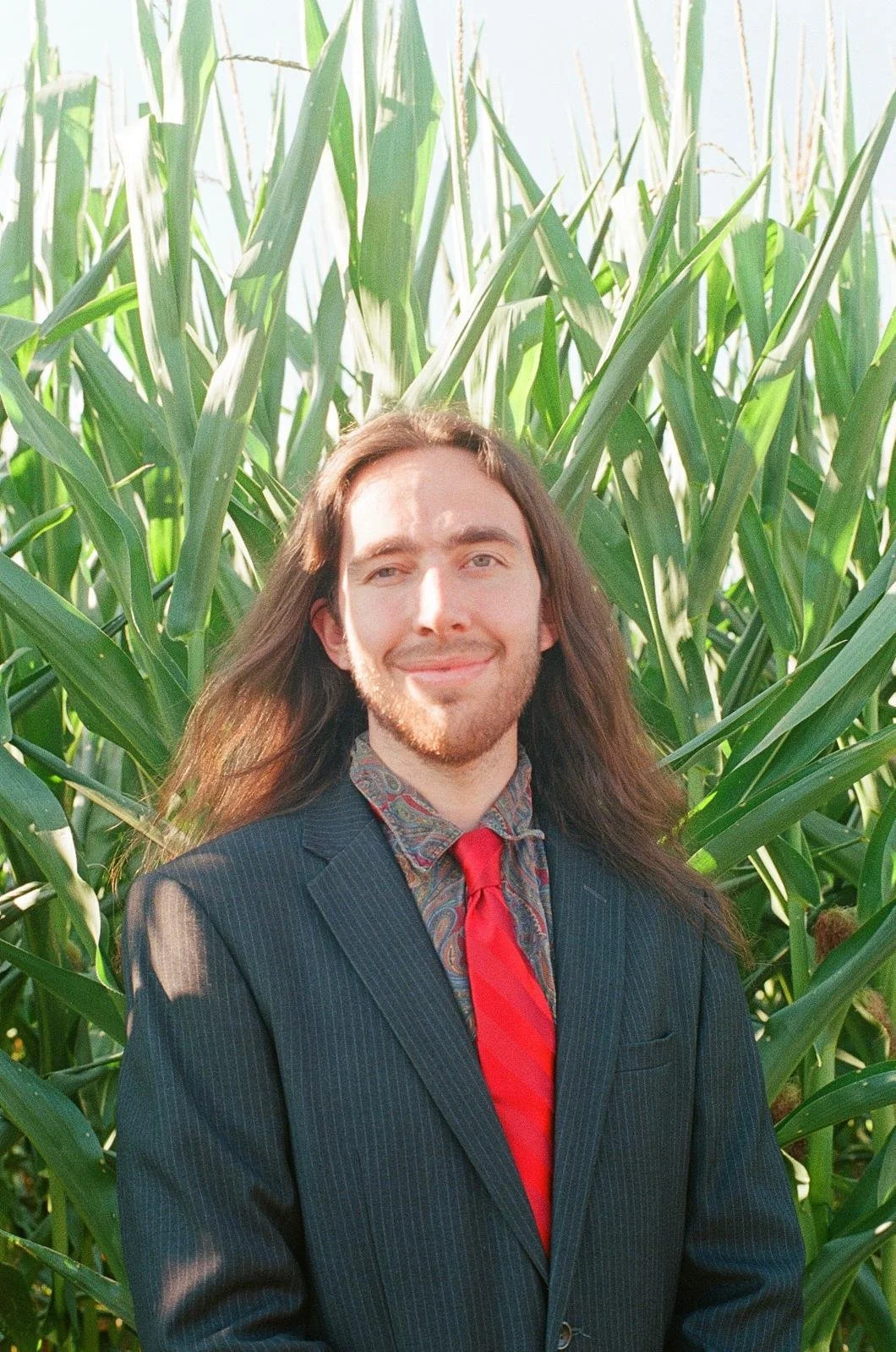 A man in a pinstripe suit, patterned shirt, and red tie standing among tall green corn stalks outdoors, smiling at the camera.